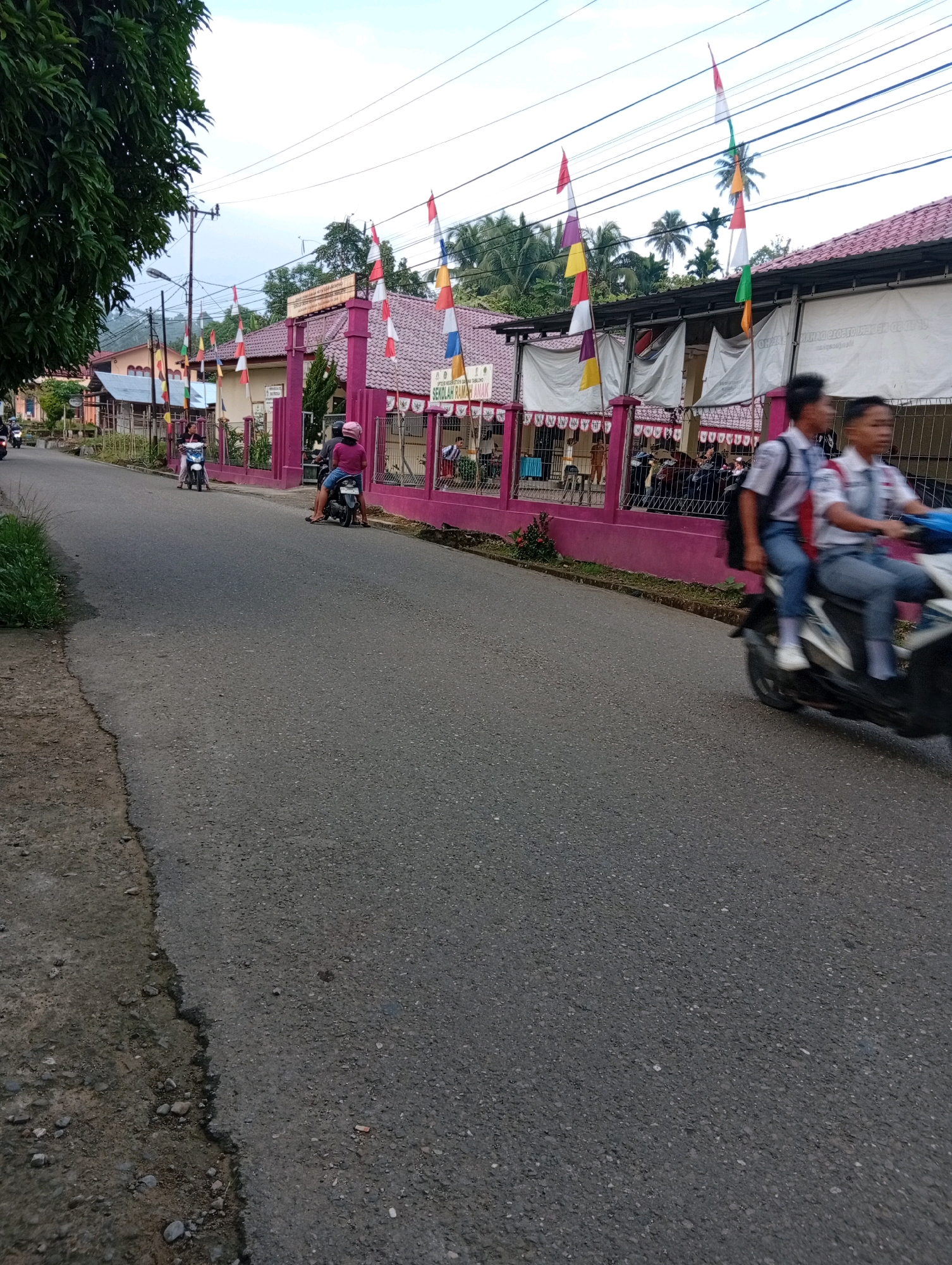 In the city of Gunungsitoli, Indonesia, a street scene unfolds in front of a school adorned with Indonesian flags and banners. A paved road runs through the foreground, with a patch of grass and dirt on the left. Several motorcycles are present on the road. Two students in school uniforms are riding a motorcycle in the middle ground, their faces blurred due to motion. Another motorcycle with a rider wearing a pink helmet is parked in the middle ground to the left of the students. In the background, the school building has a pink facade and a red tiled roof. The school is decorated with multiple red and white Indonesian flags on poles and banners. The sky is overcast, suggesting it might be late afternoon or early morning.
