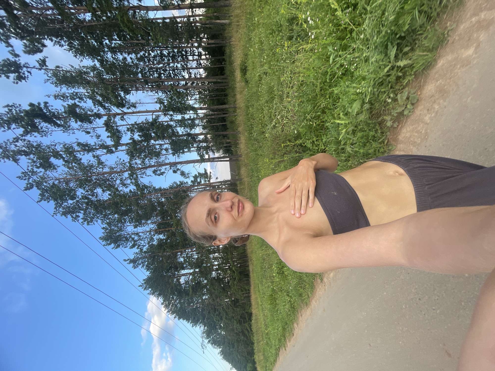 The image shows a young woman standing on a dirt road, outdoors, in a rural area. She is wearing a dark gray tube top and dark gray sweatpants. Her left hand is placed on her chest. The background consists of a field of green grass and wildflowers, with a line of tall pine trees behind it. Overhead, a bright blue sky with some wispy clouds is visible, along with power lines. The scene appears to be in a natural, undeveloped setting, consistent with the outskirts of a village or rural community like Подшивалово, Russia.