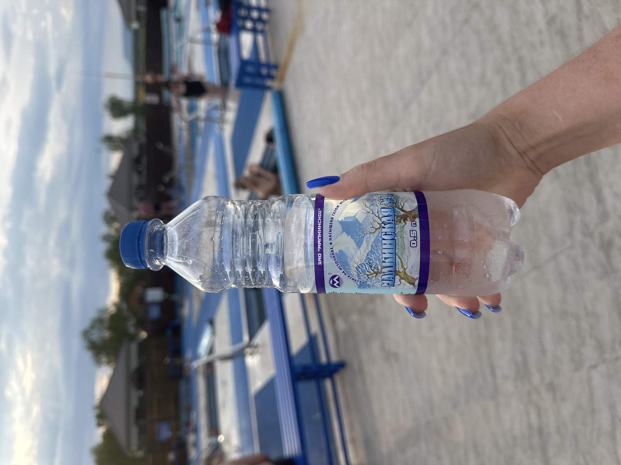 A person's hand with bright blue fingernails holds a clear plastic bottle of mineral water in front of a blurred background of an outdoor swimming pool area. The bottle has a label with text in Russian, featuring a winter landscape with mountains and a tree. The water level is about halfway down the bottle, and condensation is visible on the exterior. The background shows swimming pools with blue and white tiling, some lounge chairs, and a few people in the water or near the pools. Structures resembling small buildings or changing rooms are visible in the distance, along with trees and a cloudy sky. The lighting suggests it is late afternoon or early evening. The location is identified as the city of Yuzhnyye Koryaki, Russia, likely at a recreational facility.