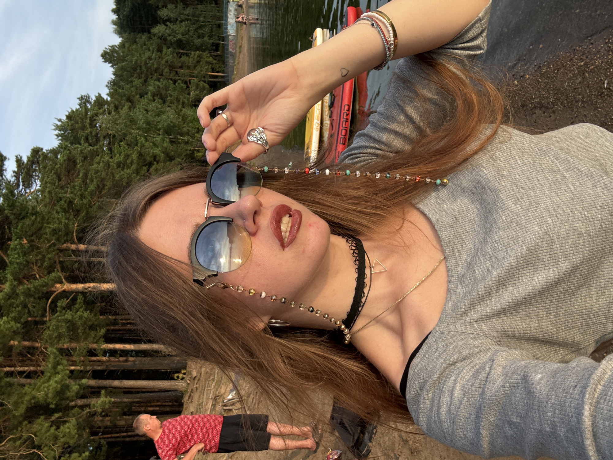 A young woman with long, auburn hair is taking a selfie outdoors. She is wearing sunglasses with a beaded chain, a choker necklace, and a grey, off-the-shoulder top. She has a bracelet on her right wrist and is wearing a ring on her left hand. In the background, a man in a red shirt and black shorts stands on a sandy shore with trees behind him. The woman is standing on the shore of a body of water with paddleboards visible. The sky is cloudy.