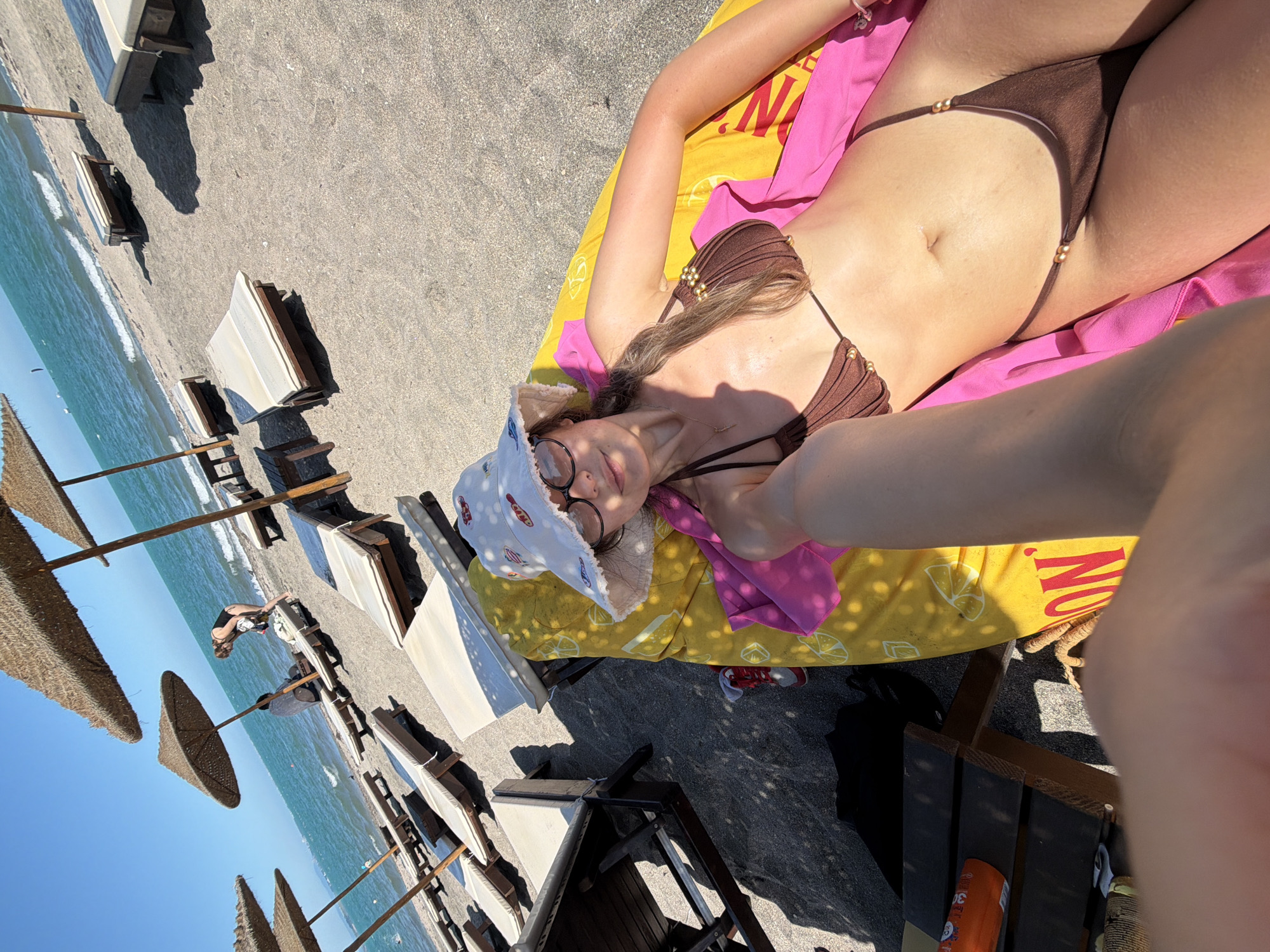 A woman is sunbathing on a beach in Burgas, Bulgaria. She is lying on a yellow beach towel, wearing a brown bikini and a white bucket hat with red designs. The beach has dark sand, many empty lounge chairs, and straw umbrellas providing shade. The sea is visible in the background with gentle waves and clear blue skies. There are a few other people in the distance, and some lounge chairs are occupied. A pink towel is draped over the woman. The overall scene depicts a sunny day at the beach.