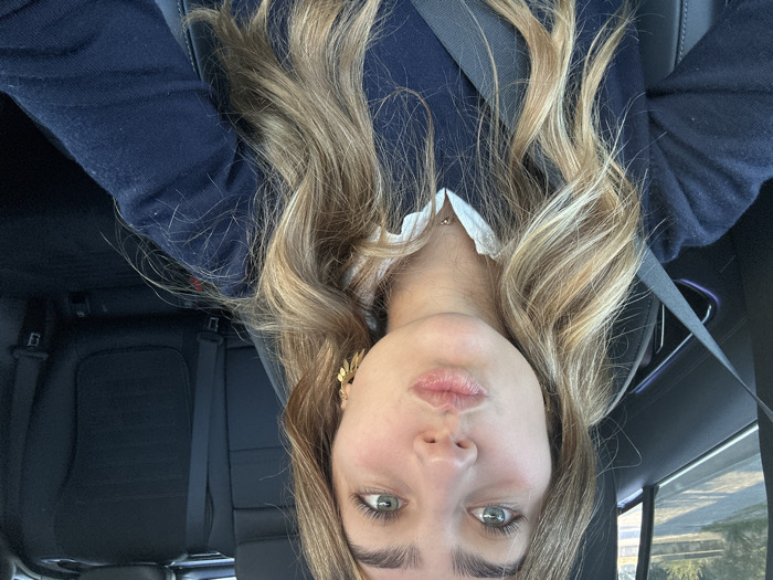 A young adult female is seated in the front passenger seat of a vehicle during daylight hours, captured in a selfie-style shot. She has fair skin, light-colored eyes, and long, wavy blonde and light brown hair. Her lips are pursed into a "kissing" gesture. She wears a dark blue V-neck sweater over a white collared shirt and a thin gold-colored necklace with a small pendant. A gold-colored earring with a leaf-like design is visible on her right ear. A dark grey seatbelt is fastened diagonally across her chest. The vehicle's interior features dark-colored seats, visible in the background rear, and dark dashboard elements. Through the window on the left, a blurred outdoor scene shows a road surface and green foliage under natural lighting. The specific location within Aldoar, Foz do Douro e Nevogilde, Portugal, cannot be determined from the visual evidence in the image.