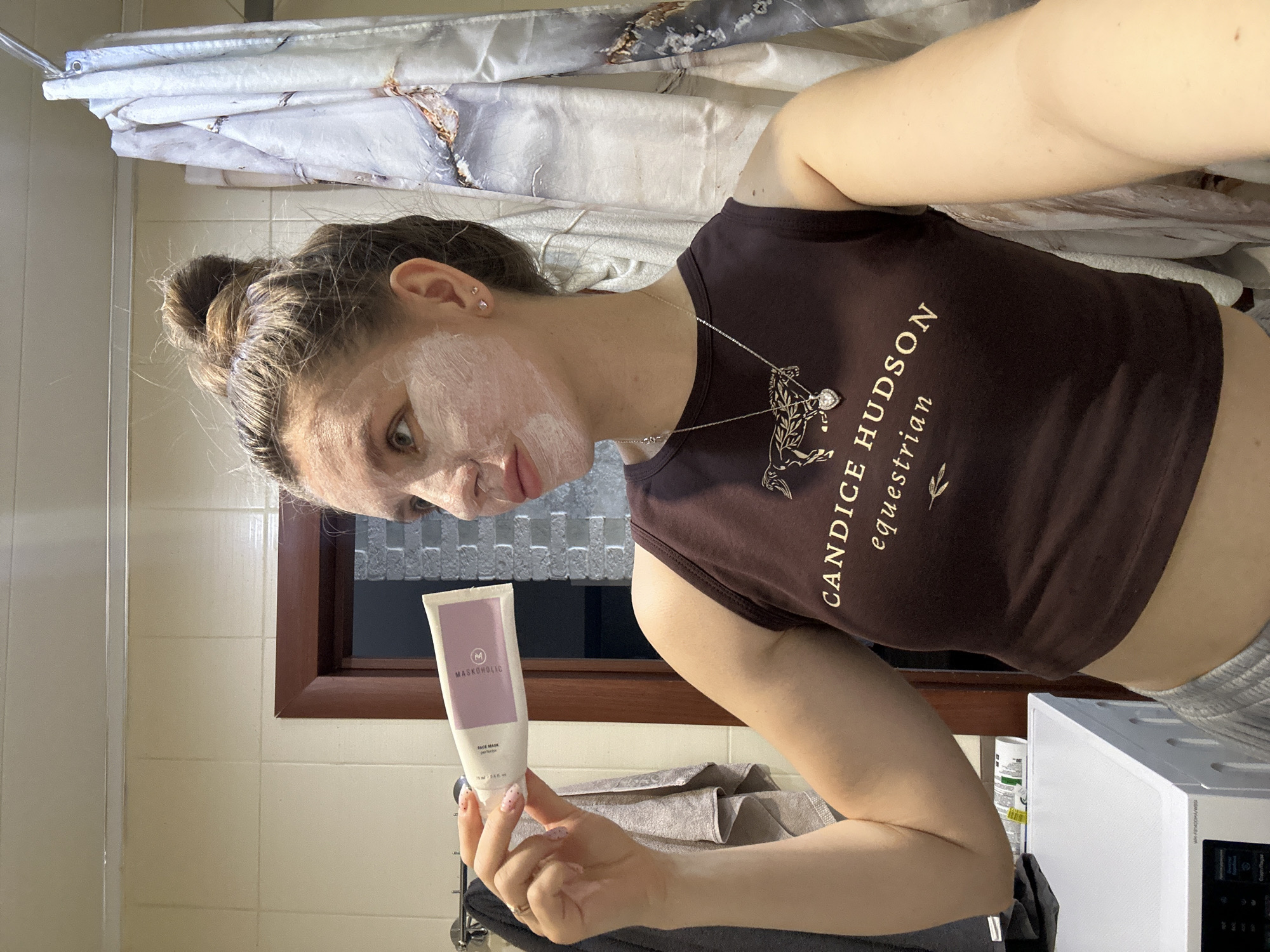 A young woman is taking a selfie in a bathroom. She has a white face mask applied and is holding a tube of "Maskoholic" face mask. She's wearing a brown cropped tank top with the words "Candice Hudson Equestrian" on it and a silver necklace with a horse pendant. Her hair is pulled back in a bun. The bathroom has white tile walls, a window with a dark frame, and a shower curtain with a marble pattern. A washing machine is visible in the background. The location is unknown.