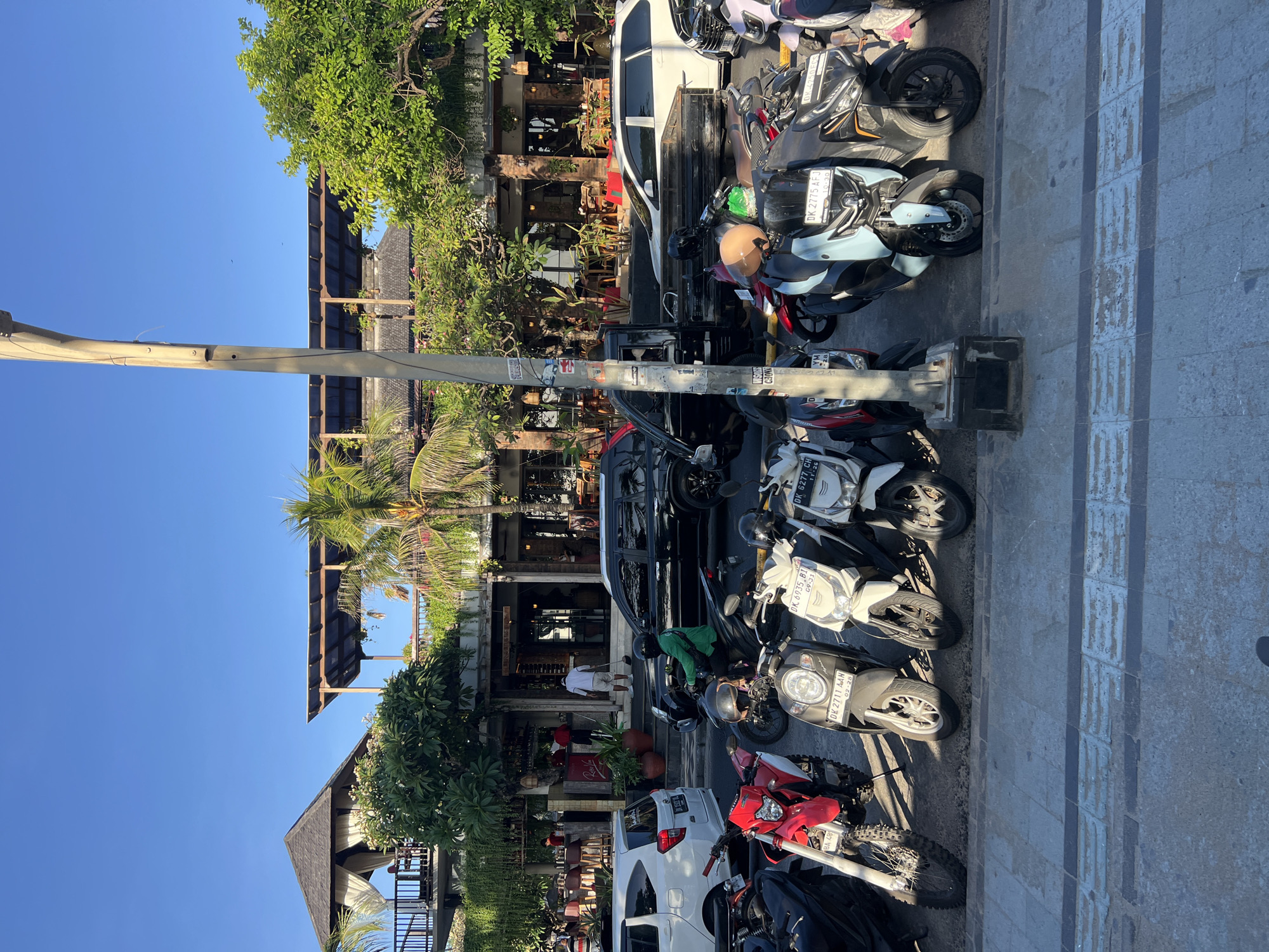 In Kuta, Indonesia, a street scene captures a row of parked motorcycles and cars in front of a multi-level building with an open-air upper level and an enclosed ground floor. The building's facade is characterized by exposed brick and wooden beams, with numerous tables and chairs set up both inside and outside, suggesting a restaurant or cafe. Palm trees and lush greenery surround the building. A person in a green shirt rides a scooter, heading away from the viewer. The street is paved with cobblestones, and a large lamp post stands prominently in the foreground. The sky is clear and blue, indicating a sunny day.
