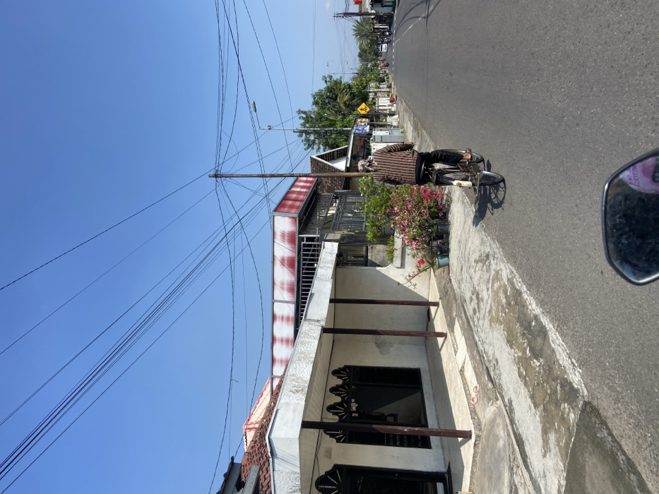 The image shows a residential street in Mojosari, Indonesia. The street is paved and lined with houses on both sides. A person is riding a bicycle down the middle of the road, heading toward the viewer. The person is wearing a checkered shirt and has a headscarf on. There are trees and bushes along the sides of the street, and numerous power lines run across the top of the image. The sky is blue with no clouds. The street is quiet and peaceful.