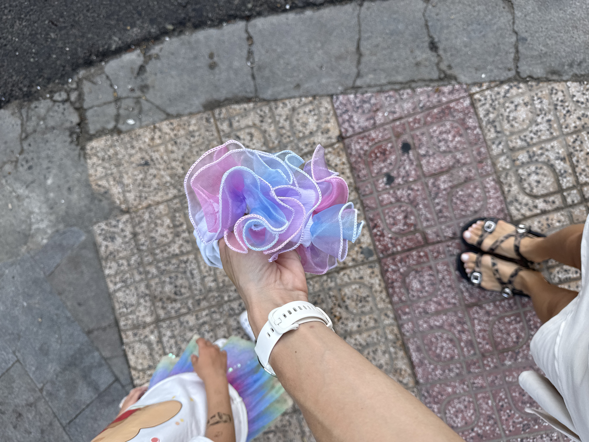 A person's arm, wearing a white smartwatch, extends into the frame holding a ruffled fabric item with pastel pink, blue, and purple hues. A portion of another person's torso and rainbow-colored skirt is visible in the upper left background. Both individuals are standing on a sidewalk composed of interlocking stone tiles and cracked gray concrete. The image is taken from a high-angle perspective.