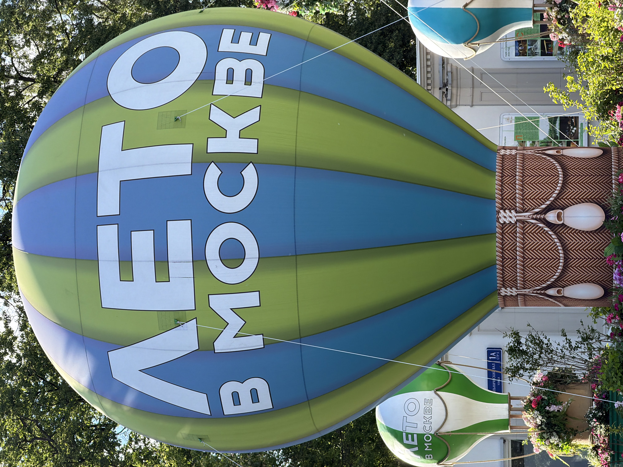 This image captures a vibrant outdoor decorative display featuring two large inflatable hot air balloons in Moscow, Russia, under bright daylight with clear skies.
The main subject is a large, colorful hot air balloon, predominantly striped in shades of blue and lime green. White Cyrillic text, reading "ЛЕТО В МОСКВЕ" (Summer in Moscow), is prominently displayed across its surface. The balloon's base is designed to resemble a woven wicker basket, with decorative elements that mimic ropes and white sandbags.
To the right and below the large balloon, a smaller inflatable hot air balloon is visible, featuring green and white stripes and the same "ЛЕТО В МОСКВЕ" text. Both balloons are held in place by numerous white strings or ropes, suggesting they are part of a fixed installation rather than actively flying.
The background features lush green trees with dense foliage, indicating a park or tree-lined street. Behind the balloons, part of a light-colored building facade is visible, adorned with flowering plants and bushes. A blue street sign on the building clearly displays "СМОЛЬНИЧЕСКИЙ ВАЛ 1А СТР 5" (Smolnicheskiy Val 1A, Building 5), providing a specific location in Moscow. Other signs on the building include text fragments like "95 лет", "2026 ГОД", and "ПАРК-СОКОЛЬНИКИ 2008 ГОД", which might refer to historical dates or anniversaries.
The overall scene suggests a festive or celebratory event, likely a summer festival or public display, given the "Summer in Moscow" theme and the decorative nature of the hot air balloons. No people are visible in the image.