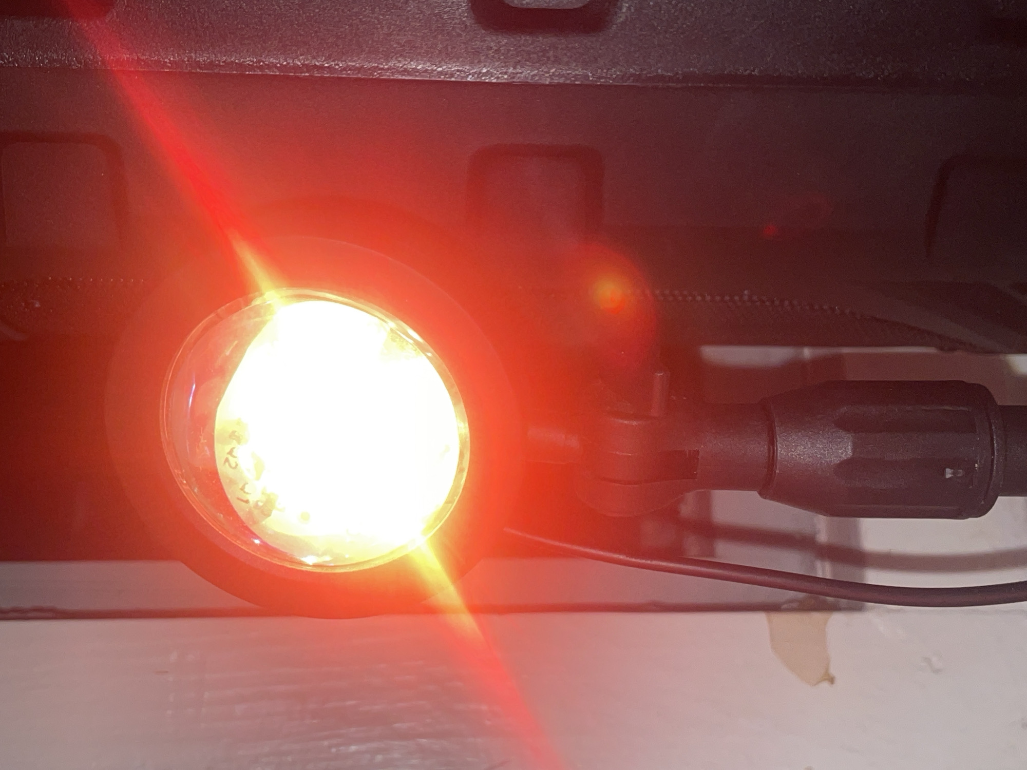 A close-up, slightly blurry, indoor shot shows a bright, white LED light source with a red halo, encased in a black circular housing. The light is mounted on a black adjustable stand, with visible textured grip points and a cable extending downwards. The background consists of a textured white wall on the left, with a light beige peeling sticker, and a dark, textured vertical object, possibly a speaker or cabinet, on the right. Red light beams emanate from the light source, casting strong red hues across the scene. The overall impression is one of a stage or studio lighting setup. The image does not contain any discernible elements that would allow for identification of its location in Saint Petersburg, Russia.