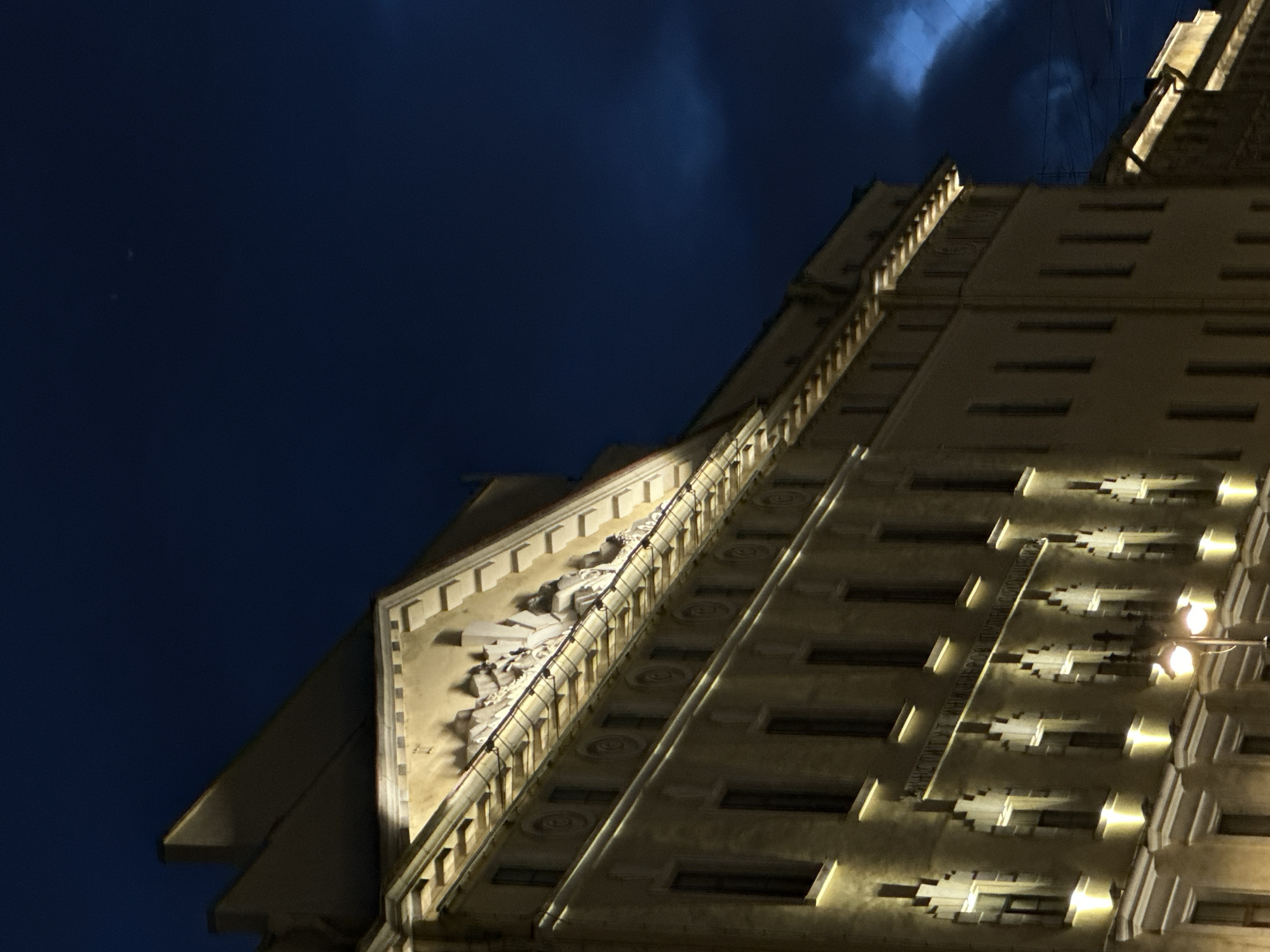An exterior, low-angle, medium shot captures the facade of an ornate, light-colored building at night. The building is illuminated by spotlights that cast a warm glow, highlighting architectural details such as a triangular pediment adorned with sculptures. A row of rectangular windows is visible on multiple levels, some with subtle light emanating from within. Below the windows, decorative elements and a lighter section of the facade can be seen. A street lamp with two lights stands in the lower-left portion of the frame, casting its own illumination. The sky above is a deep, dark blue, with a hint of lighter, cloud-like formations on the right side. The overall impression is one of grand architecture under a night sky.