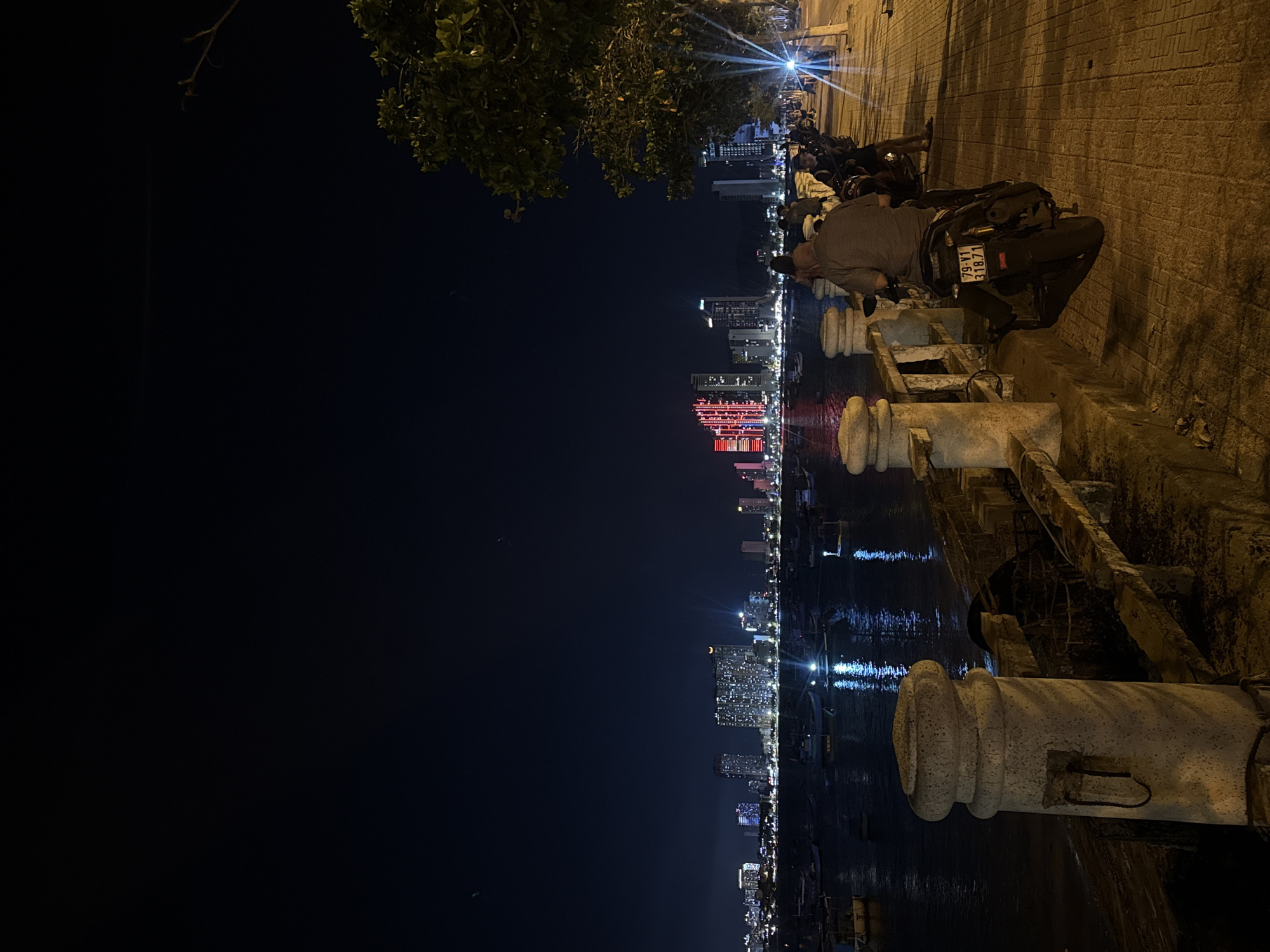 The image depicts a nighttime scene in Bac Nha Trang Ward, Vietnam, likely along the waterfront. In the foreground, a paved walkway runs alongside a stone barrier overlooking a body of water. Several motorcycles are parked along the walkway, and two individuals are seated on one of them, facing away from the camera. One person is wearing a grey shirt and the other is partially obscured.
Beyond the walkway and water, a city skyline is visible, illuminated by the lights of numerous buildings. Some buildings are brightly lit with white lights, while a prominent cluster of skyscrapers in the center is illuminated with red lights. A fleet of boats is anchored in the water, with their lights reflecting on the dark surface. The sky above is a deep, dark blue, with a few faint stars or distant lights visible. The overall atmosphere is urban and nocturnal, with the city lights creating a vibrant backdrop to the more subdued foreground.