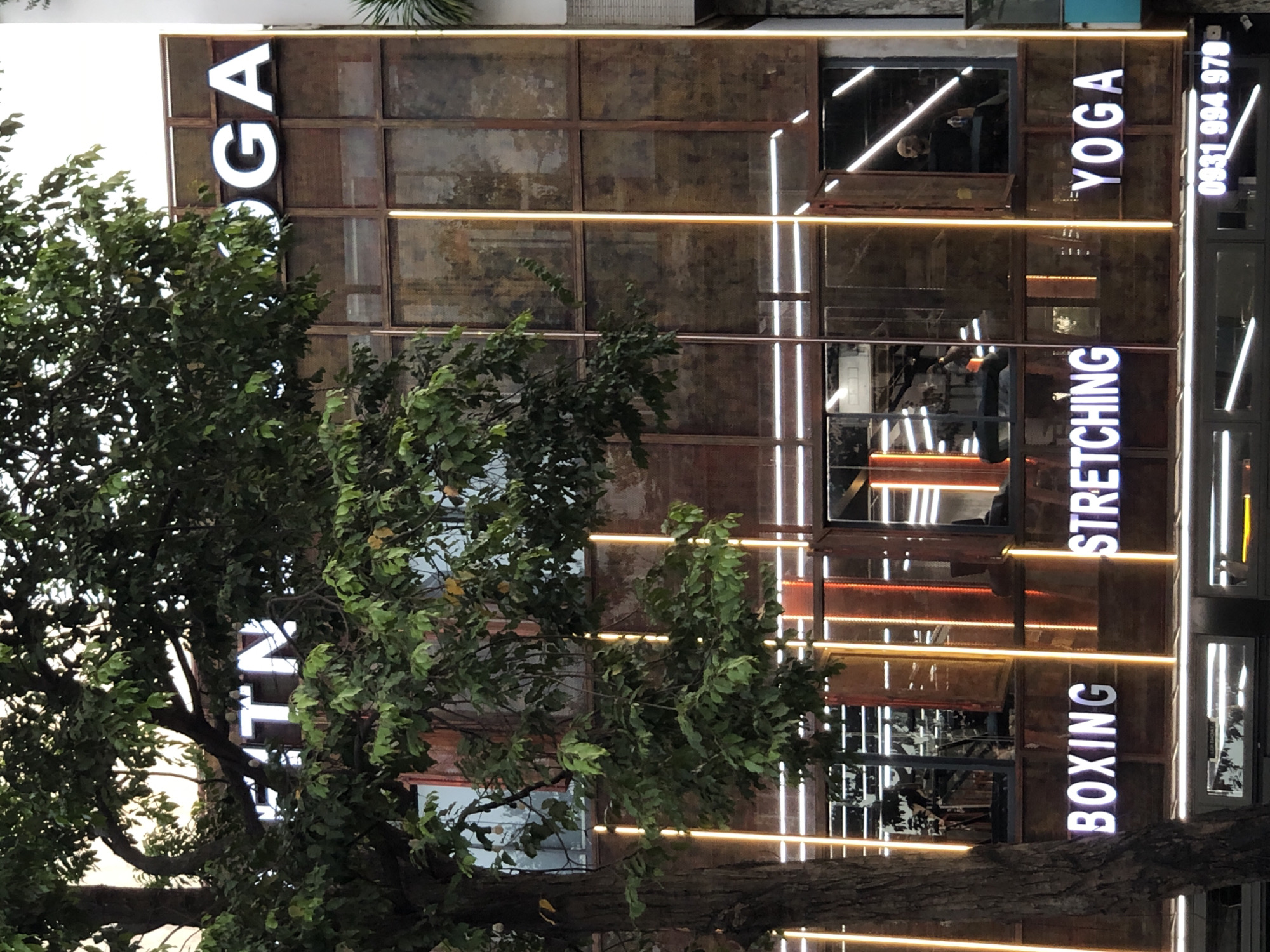 The image shows the facade of a fitness center in Da Nang, Vietnam. The building has a modern industrial design with large glass windows and rustic metal accents. The words "FITNESS", "BOXING", "STRETCHING", and "YOGA" are prominently displayed in large, illuminated letters across the facade. Vertical orange and white lights are installed along the building, accentuating its structure. Inside the windows, fitness equipment and people can be seen. A tree with dense foliage partially obscures the view of the building. The lower part of the building features a storefront with a phone number displayed.