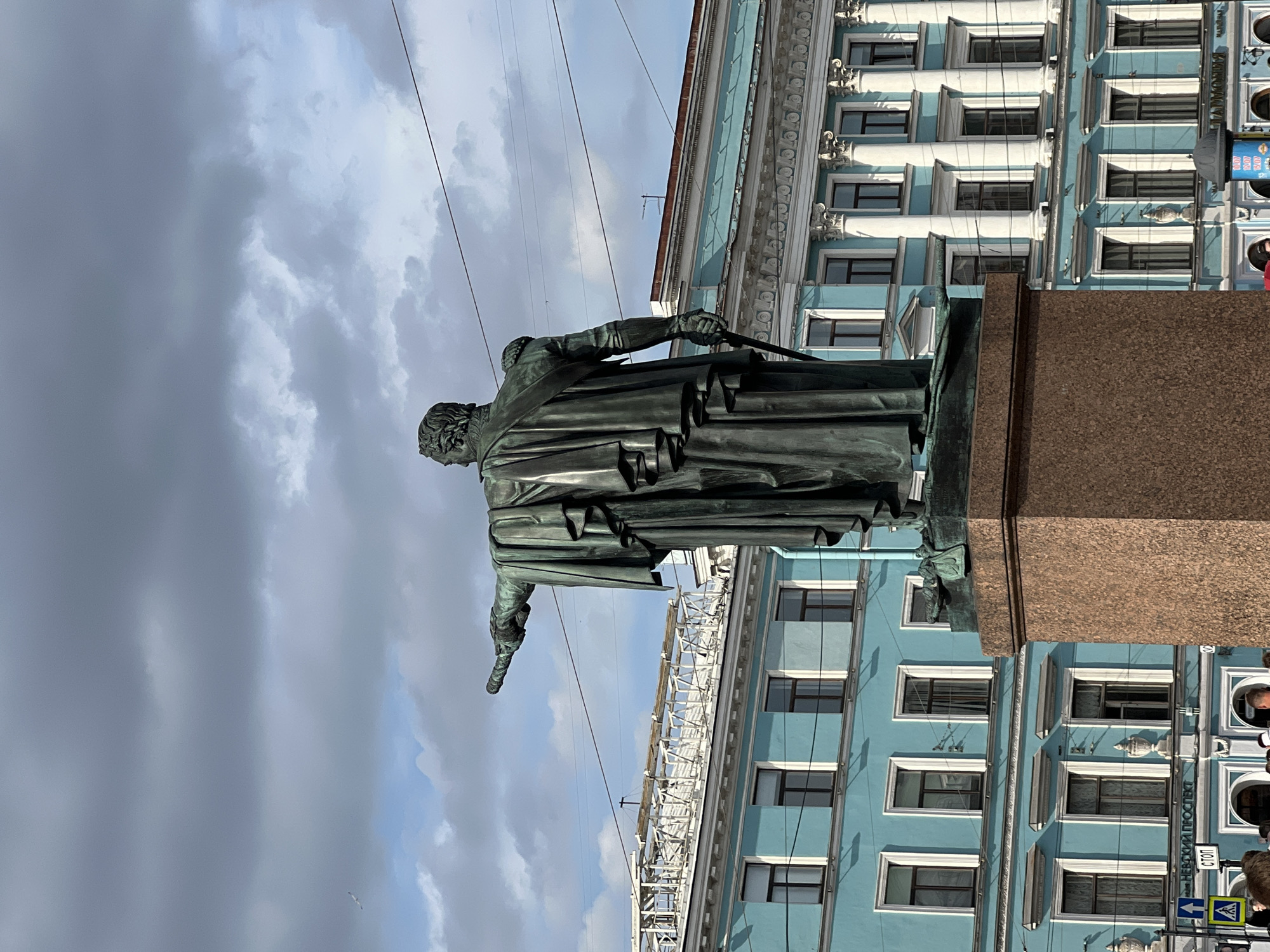 This is a photograph taken outdoors in Saint Petersburg, Russia. The main subject is a bronze statue of a man, viewed from behind and looking to the left. He is dressed in a long coat and appears to be holding a sword or similar object. The statue stands on a large, reddish-brown granite pedestal.
In the background, a light blue building with white trim and numerous windows rises behind the statue. The building has decorative architectural elements, including columns and molding. Scaffolding is visible on a section of the building, suggesting construction or renovation. Power lines stretch across the sky in front of the building.
The sky is filled with dramatic, heavy grey clouds, with patches of brighter sky peeking through. The lighting suggests it is daytime, possibly late afternoon or early evening, given the somewhat subdued light.
Visible text on a sign on the blue building reads "НЕВСКИЙ ПРОСПЕКТ" (Nevsky Prospekt), a famous street in Saint Petersburg, and "СТОП" (Stop). There are also traffic signs with yellow and black markings. The presence of these signs and the architecture strongly indicate an urban setting. The overall mood is somewhat somber due to the cloudy sky.
