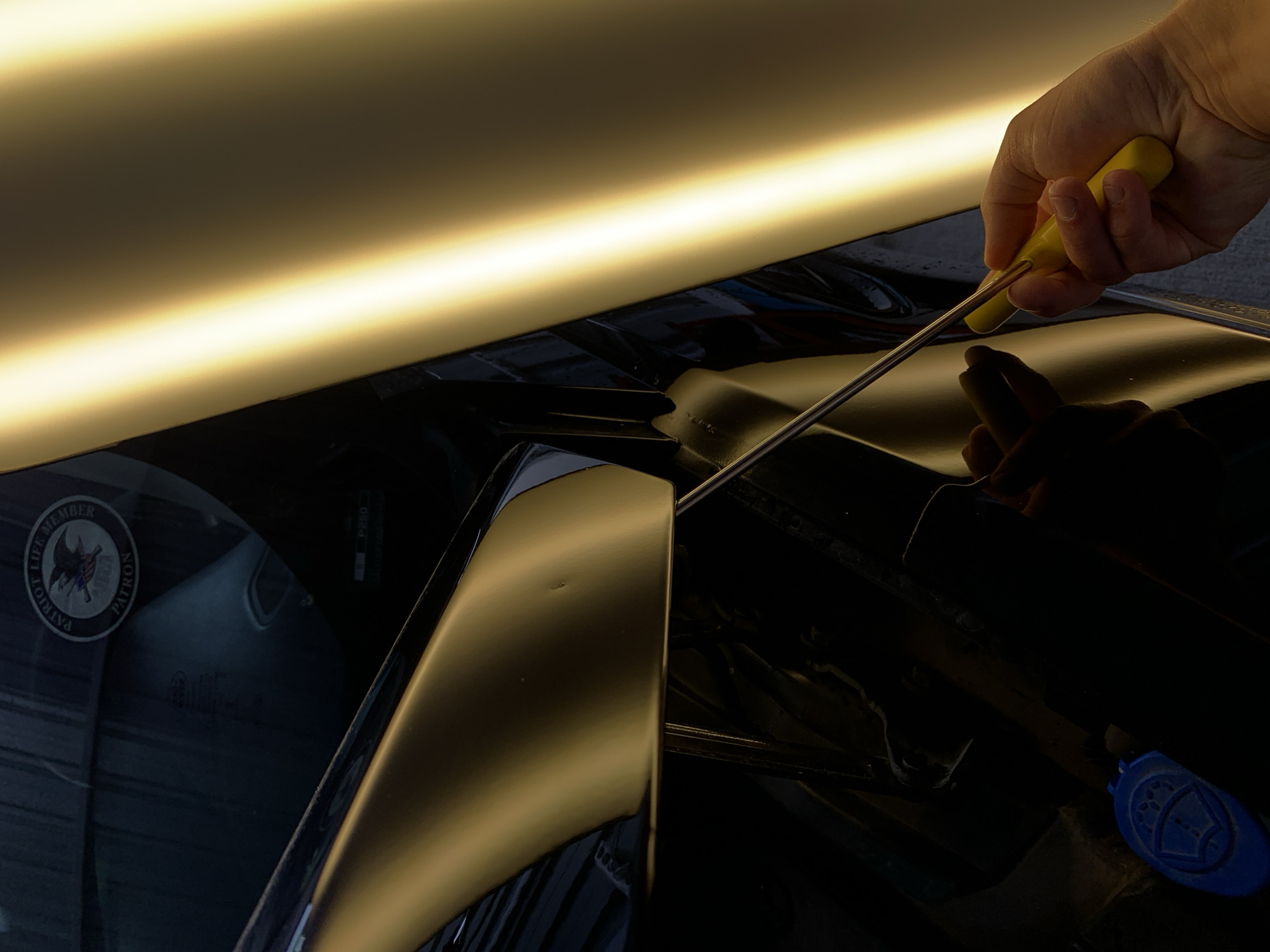A close-up shot shows a hand holding a yellow-handled tool, likely a dent repair rod, on the side of a dark-colored vehicle with a matte gold panel. The tool appears to be inserted behind the gold panel, suggesting an attempt to repair a visible dent. In the upper left corner of the image, a circular sticker with an eagle and the text "PATRIOT LIFE MEMBER" and "PATRON" is affixed to the vehicle's surface. A bright, illuminated panel runs vertically along the right side of the frame, casting a strong yellow light and creating reflections on the car's surface. The background is dark and somewhat indistinct, showing the interior of a garage or workshop. The presence of the dent repair tool and the lighting suggest this image was taken in a professional auto body shop.