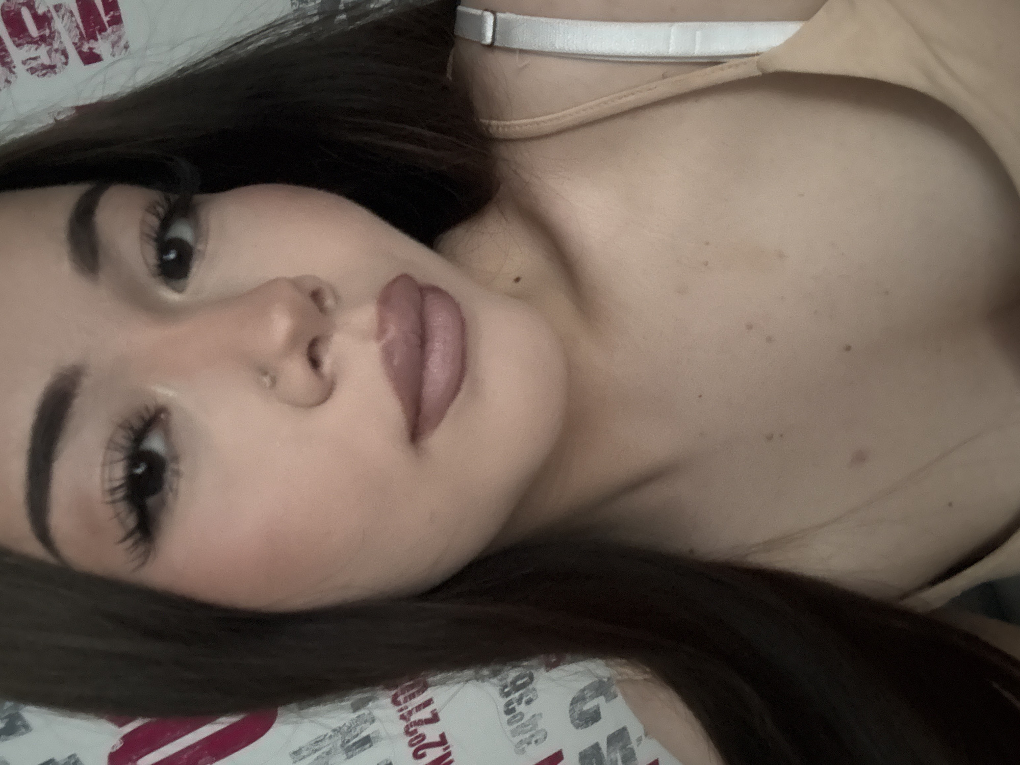 The image depicts a close-up, head-and-shoulders portrait of a young woman with long, dark brown hair, looking directly at the viewer. Her skin tone is fair to medium, and she has dark, defined eyebrows, dark brown eyes with prominent false eyelashes, and a small silver stud piercing in her left nostril. Her lips are full and coated in a matte, mauve-toned lipstick. Visible on her neck and upper chest are several small moles or freckles. She is wearing a light-colored (nude or beige) camisole or bra, with a thin strap visible over her right shoulder and a wider, light-colored strap visible on the far right of the frame. The background is a white surface, possibly a wall or fabric, patterned with large, stylized text and numbers in red and black, including partial readings of "2°21'" and "34°36'". The scene is located in Naberezhnye Chelny, Russia.