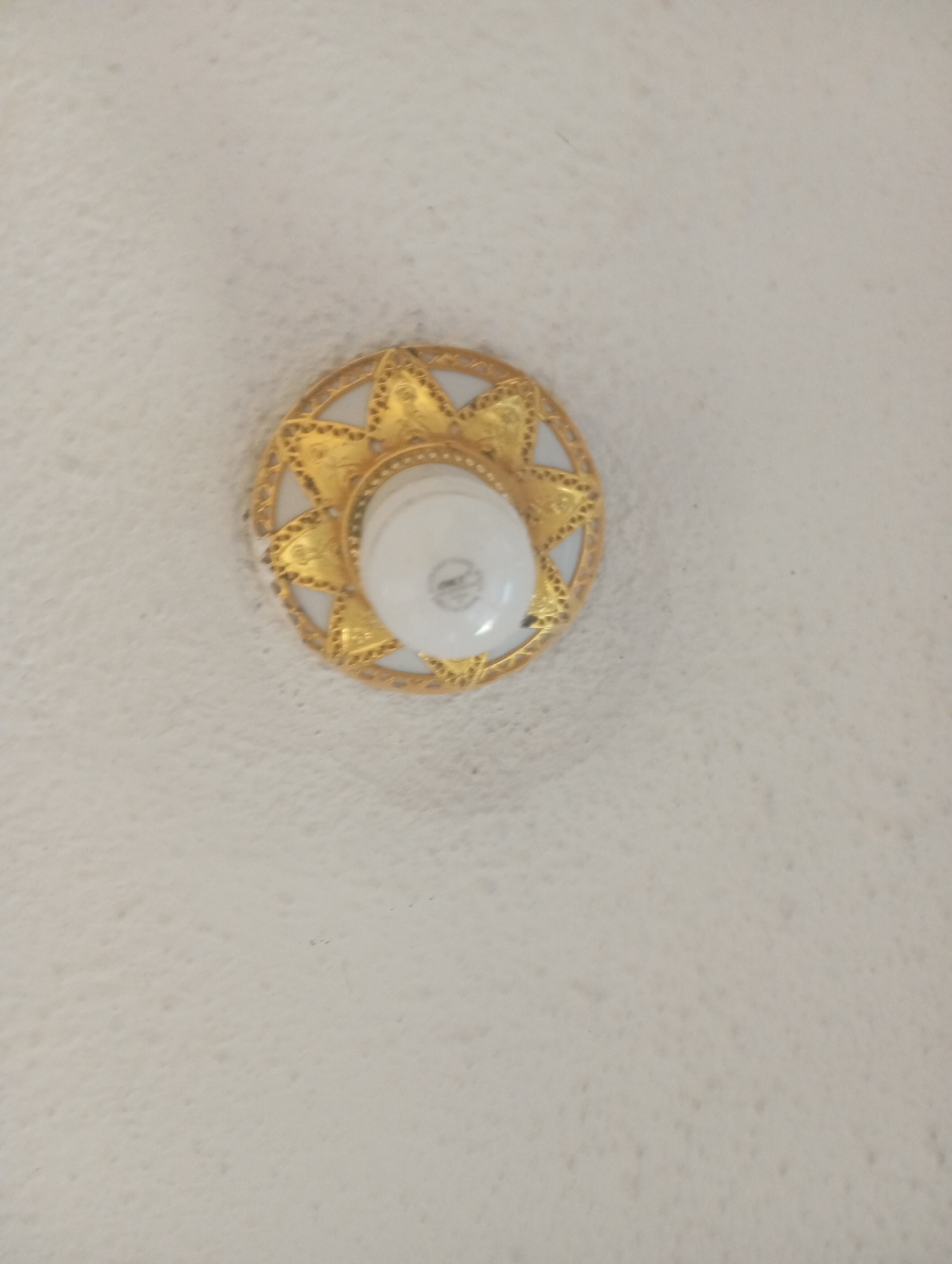 A close-up, high-angle shot shows a decorative gold-colored light fixture with a white light bulb in the center, mounted on a textured, off-white ceiling. The fixture has a circular base with a starburst pattern radiating outwards, featuring intricate cutouts and a beaded edge. The light bulb is slightly tilted and appears to be an incandescent bulb. The ceiling has a slightly uneven, stucco-like texture, with small shadows and subtle variations in tone across its surface. The image appears to be taken indoors.