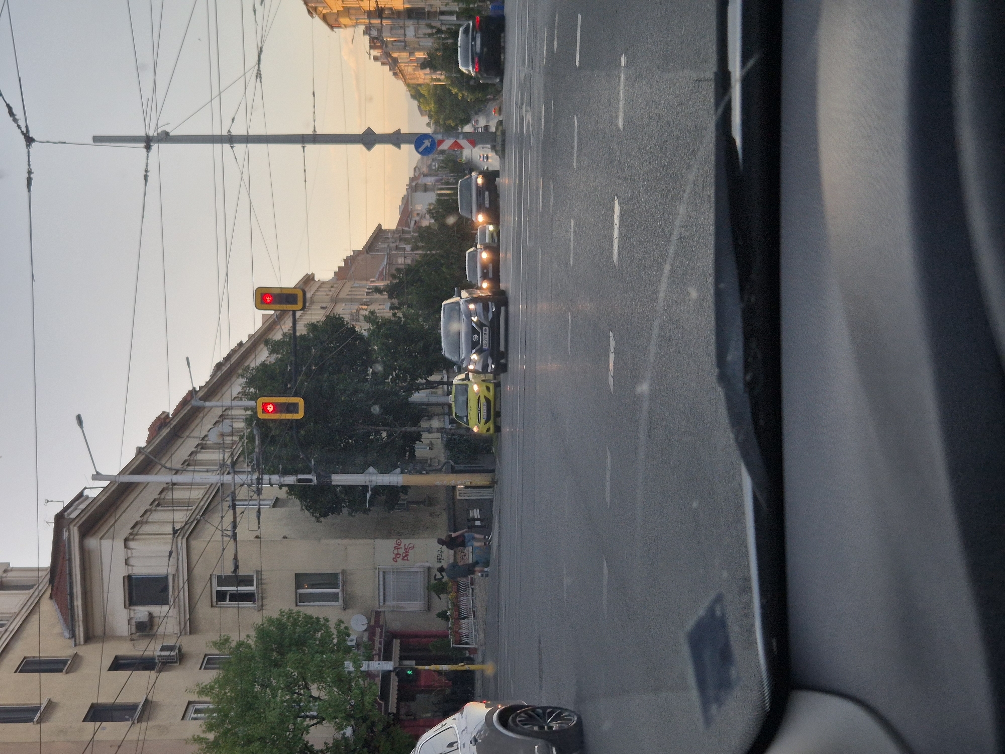 The image depicts a street scene in Sofia, Bulgaria, at an intersection. A traffic light is visible, illuminated with a red signal. Several cars are present, including a yellow taxi, a silver SUV, and other dark-colored sedans. Pedestrians are walking on the sidewalk. The architecture of the buildings lining the street suggests a European urban setting, with multi-story buildings featuring balconies and ornate details. The sky is overcast, indicating a likely overcast day or early evening. The perspective suggests the photo was taken from inside a vehicle, looking forward through the windshield. The overall impression is a typical urban street with vehicular and pedestrian traffic.