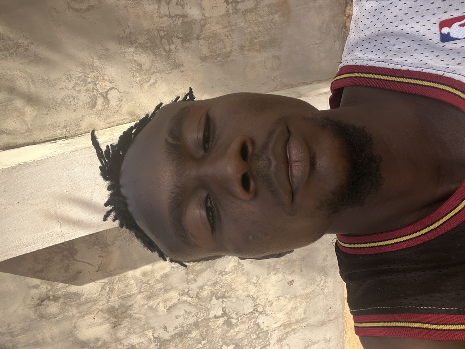 An adult male is depicted in a close-up, head-and-shoulders portrait, positioned against a textured wall in Birnin Kebbi, Nigeria. The subject has dark skin, short dark hair styled in braids or dreadlocks with faded sides, and a small goatee. He wears a basketball jersey with a split design; the left side (viewer's left) is predominantly dark brown with red and gold stripes, while the right side is white mesh featuring a red and blue NBA logo. His eyes are dark and appear slightly narrowed as he looks directly into the camera. The background consists of a light-colored, weathered concrete or plaster wall exhibiting discoloration and rough surface texture. A distinct shadow line from a vertical structural element (pillar or corner) is visible on the wall behind the subject's head. The scene is illuminated by bright natural light, casting clear shadows.