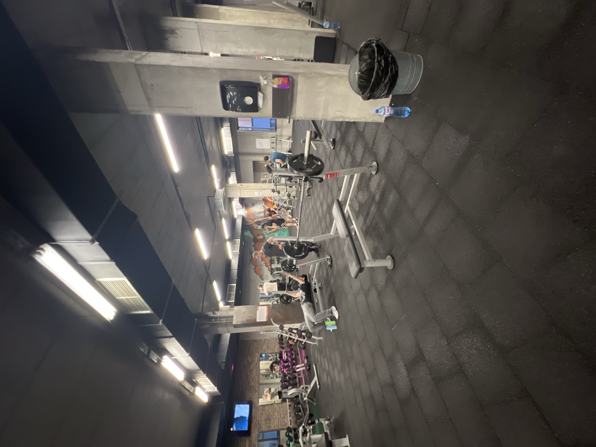 The image depicts a gym in Sofia, Bulgaria. The gym has weightlifting machines and equipment. People are exercising on the equipment. There is a trash can and a water bottle on the floor near one of the weightlifting machines. The gym has fluorescent lighting.