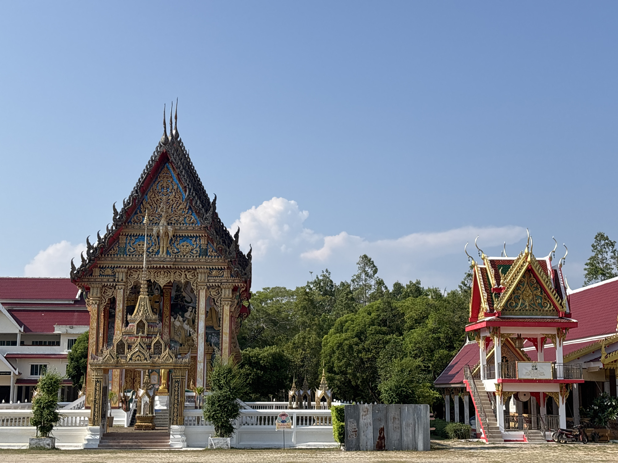 This wide-angle outdoor shot captures a vibrant temple complex in Choeng Thale, Thailand, under a clear, bright blue sky with scattered white clouds. The scene is dominated by two ornate structures set against a backdrop of lush green trees and a distant white building with a red roof.
On the left, a large, intricately decorated main temple building stands prominently. Its multi-tiered roof features dark, spiky finials and is richly adorned with gold, red, and blue decorative elements and carvings. The facade beneath the roof is equally elaborate, showcasing golden relief work, murals, and statues within alcoves. A smaller, golden tiered structure with multiple pillars is positioned at the entrance of the main temple. In front of this building, there's a white fence or wall, flanked by small potted green shrubs. A white sign near the fence on the left reads "บุญรักษา" (Bun Rak Sa), meaning "May merit protect you" or "Blessing" in Thai.
To the right, a smaller bell tower building features a distinctive red roof with golden trim and gables, characteristic of traditional Thai architecture. This structure has a white base and a staircase leading up to an open-air platform, where a large bell or drum is likely housed. A white plaque with Thai script and what appear to be two small portraits is affixed to the platform's railing. A motorbike is parked near the base of the bell tower, and a corrugated metal fence borders the right edge of the frame.
The overall impression is one of cultural richness and architectural splendor, set within a serene, sunny environment. No people are visible, suggesting a tranquil moment.