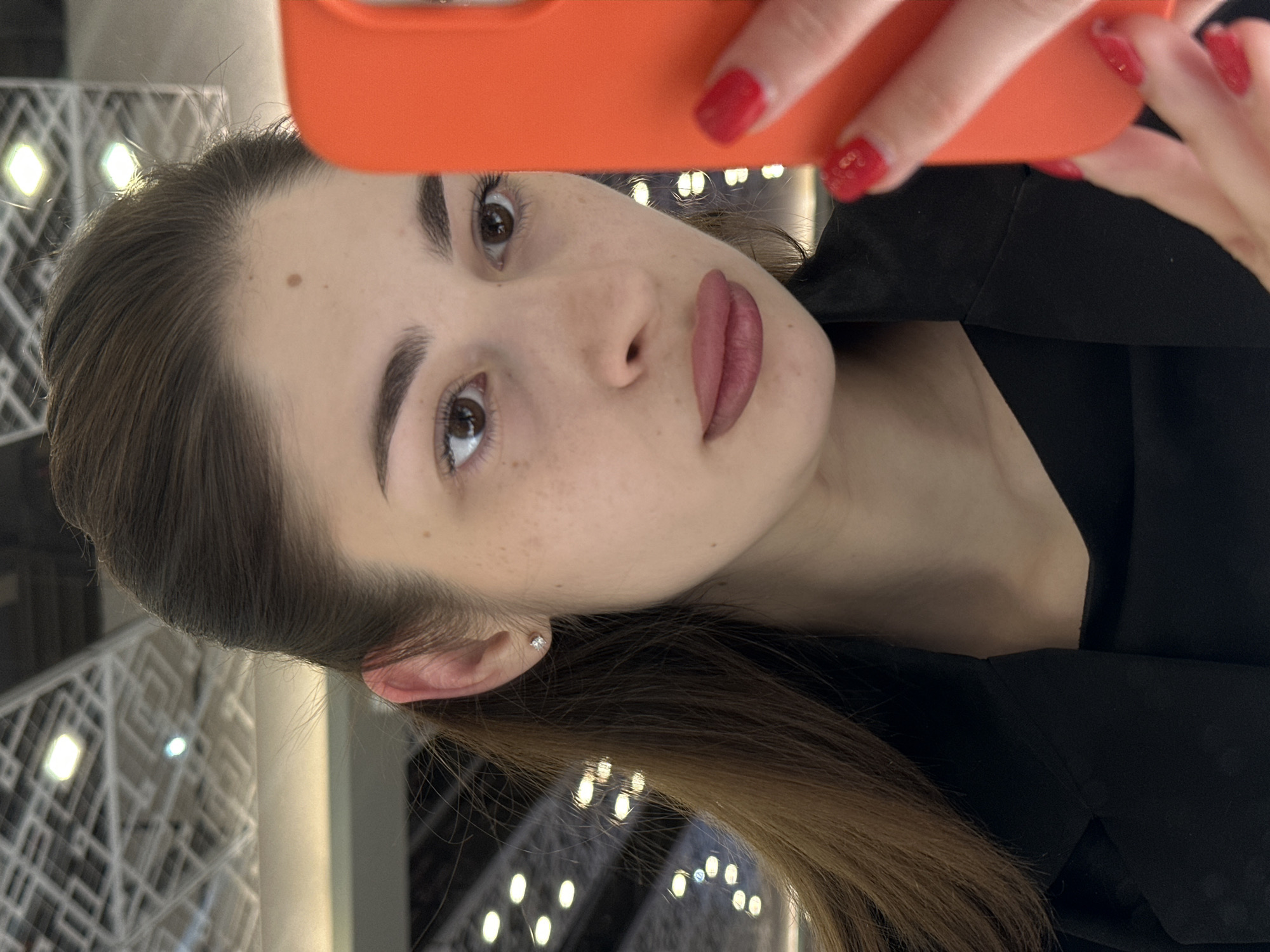 A female subject, appearing to be an adult, is depicted in an indoor setting in Yekaterinburg, Russia. She has brown hair, styled partly pulled back, with longer strands cascading over her left shoulder. Her skin tone is fair with visible freckles on her cheeks. A prominent mole is located on her forehead, slightly right of center. She has brown eyes, which are directed downwards towards a bright orange smartphone held in both hands. Her lips are adorned with a matte, plum-toned lipstick. She wears a small, clear stud earring in her left earlobe. Her attire consists of a black top or undershirt, partially visible beneath a black blazer or jacket. Both of her hands, with nails painted bright red and featuring small decorative accents, are holding the orange smartphone, which obscures the right side of her face. The background, out of focus, reveals a light-colored, geometrically patterned ceiling with multiple rectangular light fixtures, indicative of a modern public building interior.