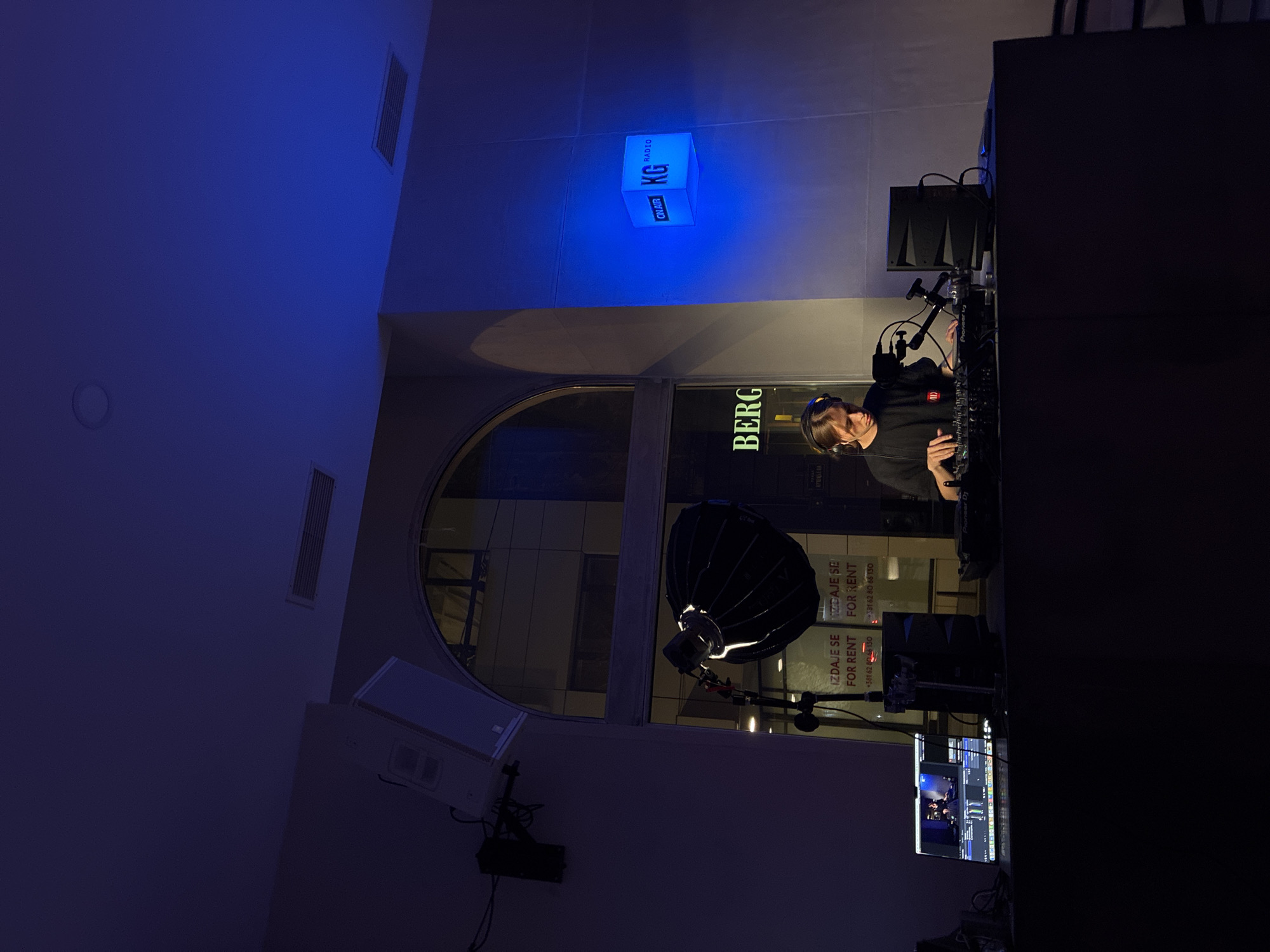 The image shows a scene in what appears to be a radio station or broadcasting studio, likely in Belgrade, Serbia, based on the "BERG" signage and the presence of Serbian text ("IZDAJE SE FOR RENT") visible in a window. A person wearing headphones is operating a DJ console, with sound equipment visible nearby. A "KG RADIO" on-air light is also visible. The room is illuminated by a combination of natural and artificial light, including blue lighting effects. A softbox light is also present. A monitor showing what appears to be a broadcast program.