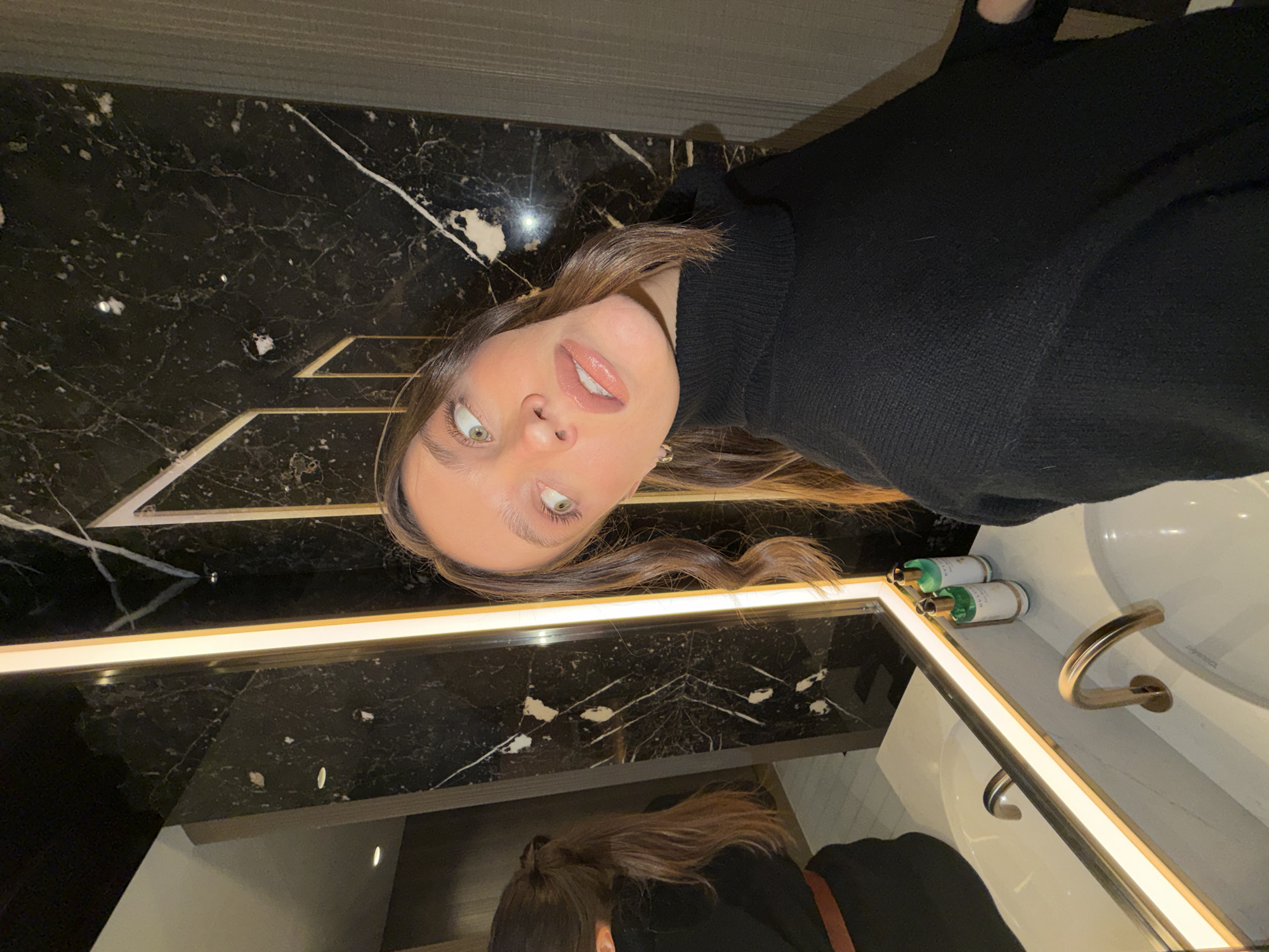 A female subject is depicted in a modern, luxurious bathroom setting, appearing to capture a mirror selfie. She has long, wavy brown hair and wears a black turtleneck sweater. Her head is tilted to the right, and her light-colored eyes are directed upwards and left. Her lips are glossy. The subject's reflection in the mirror shows her back, with her hair in a ponytail, wearing a black garment and a brown belt. The bathroom features dark marble walls with prominent white veining and geometric gold-colored decorative accents. A large mirror with an integrated vertical LED strip light is positioned above a white sink, which has a distinctive gold-colored arched faucet (Duravit brand visible). Two bottles of toiletries, one identifiable as BVLGARI, rest on the countertop. The precise location within Budva, Montenegro, is not discernible from the visual information, but the upscale design suggests a high-end hotel or private residence.