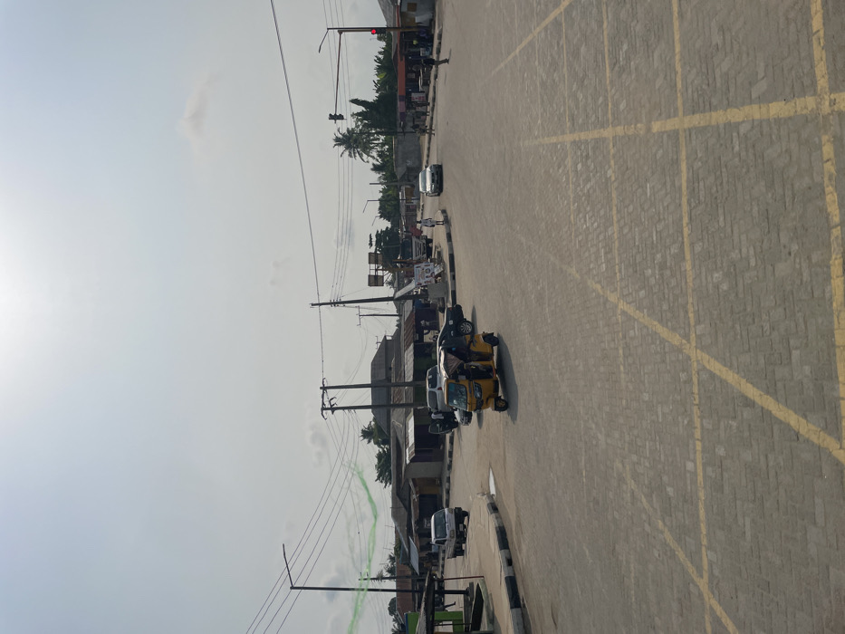 This image depicts a bustling intersection in Mosafejaw, Nigeria, under a bright, hazy sky. The foreground shows a grey interlocking brick road surface with prominent yellow cross-hatching, indicating a no-stopping zone.
Traffic is present, featuring a yellow motorized tricycle (keke napep) in the mid-ground, heading right, with at least two occupants. Behind it, a dark sedan is parked or stopped with its driver's side door open, adjacent to a white van also with an open door. Further left, a white pickup truck is partially visible, moving away from the viewer. In the distance, a white sedan approaches the intersection from the right. A large white truck is partially visible behind utility poles in the mid-ground.
Several pedestrians are visible; one person in a white top walks near the approaching white sedan, and two others are on the far right side of the road.
The intersection is lined with single-story buildings, many featuring corrugated metal roofs, indicative of commercial or residential structures. Dense utility poles with numerous overhead power lines crisscross the scene. A red traffic light is visible on the far right, and a road sign structure stands in the mid-ground. Palm trees and other tropical foliage are evident in the background, particularly on the right. A faint, irregular green line or streamer is visible in the upper left sky.