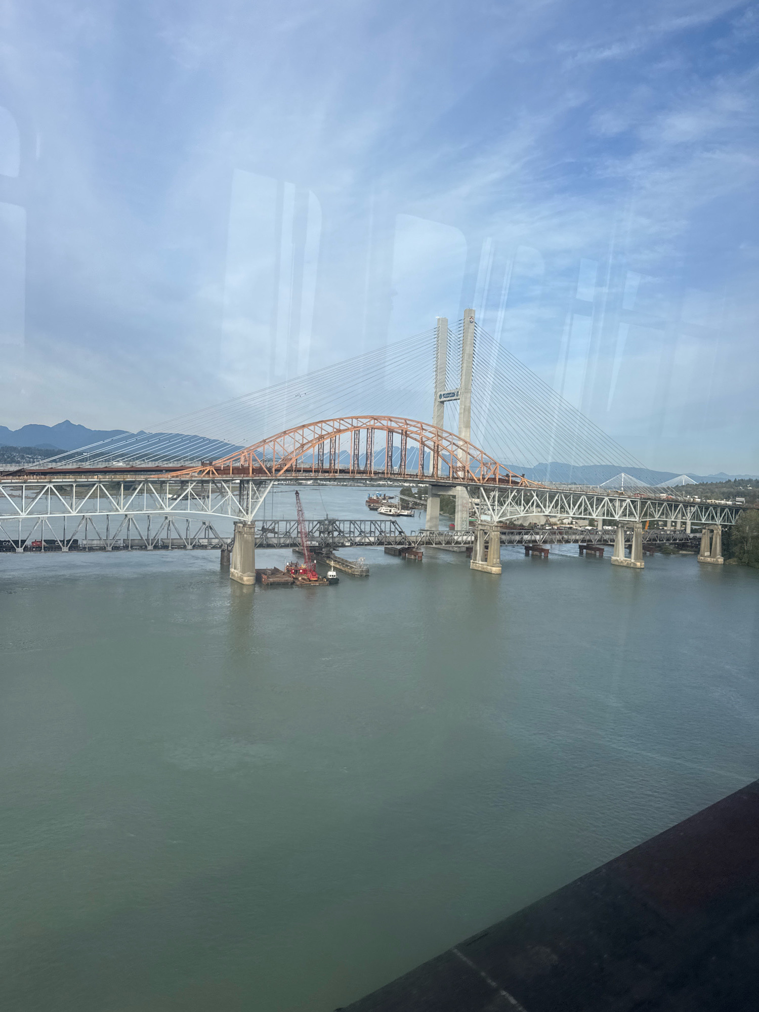 The image depicts a wide view of the Fraser River in New Westminster, Canada, featuring a complex of bridges. The foreground is dominated by the murky, greenish water of the river, with a dark, out-of-focus element along the bottom right edge, possibly a railing or part of the vantage point.
Dominating the scene are multiple bridges spanning the river. The most prominent is a large cable-stayed bridge with two distinctive white towers, its cables fanning out over the water. Adjacent to this, and slightly closer, is an older-style bridge with a distinct orange arch, appearing to be a truss bridge. Further to the left, another multi-span bridge with white metal girders and concrete piers is visible, suggesting a rail bridge or a different road bridge.
In the water below the bridges, several barges and construction vessels are present. Red cranes are visible on some of the barges, indicating ongoing construction or maintenance work. A small white tugboat is also positioned near the construction barges.
The background reveals a hazy mountain range in the distance, beneath a pale blue sky with scattered white clouds. The overall impression is a busy urban waterway with significant infrastructure. The reflections on the glass from which the photo was taken are noticeable, particularly in the sky area. No people are clearly discernible in the image.