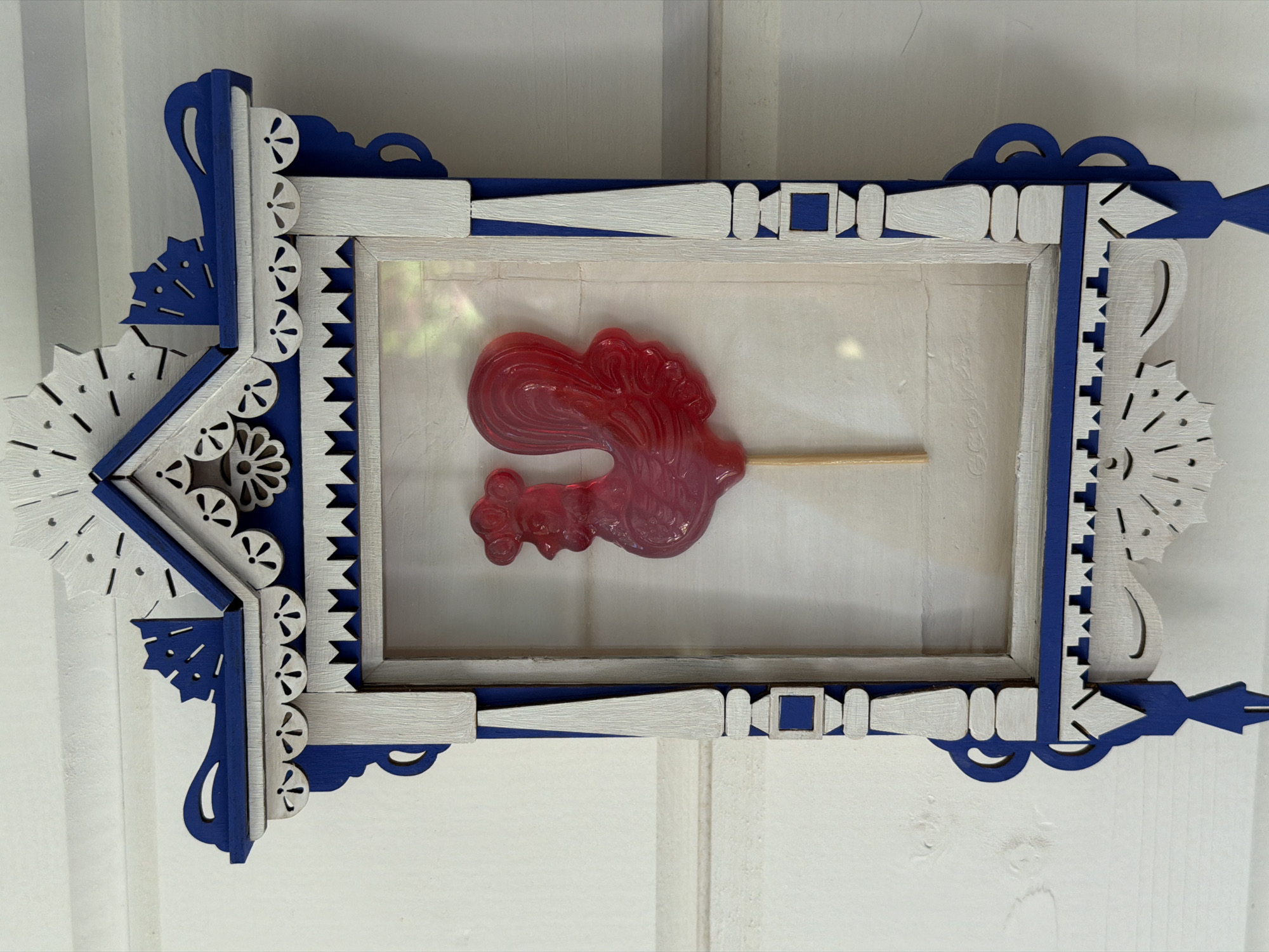 A red rooster-shaped lollipop is displayed in a carved wooden frame. The frame is painted white and blue with intricate, decorative cutouts along its edges, resembling traditional Russian folk art. The frame is mounted on a white wooden wall, with horizontal planks visible. The lollipop has a glossy finish and is attached to a light-colored stick. Sunlight casts a faint rainbow-like reflection on the back surface of the frame. The overall scene suggests a decorative display, possibly in a home or cultural setting in Алешкино Дп, Russia.