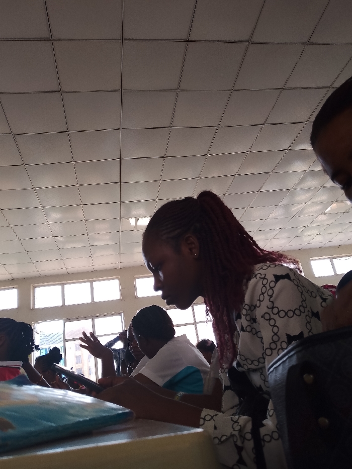 The image depicts an indoor scene, possibly a classroom or lecture hall in Umueshi, Nigeria. Several people are present, primarily female, seated at desks or tables. One woman in the foreground has her hair styled in braids and is wearing a patterned shirt. She appears to be looking down at something on the desk. Other individuals are visible in the background, with one person raising their hand. The room is well-lit by windows along the back wall, and the ceiling is tiled. A book is laying on the desk in the lower left corner. A dark bag is on the lower right. The setting suggests an educational environment, likely filled with students engaged in a class or study session.