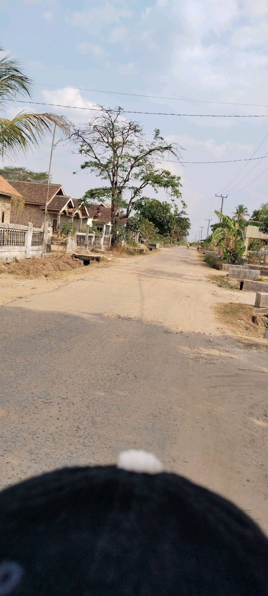 The image shows a dusty road in a rural area, likely in the village of Tanjung Ratu Ilir, Indonesia. The road is lined with houses and trees, and power lines are visible in the sky. In the foreground, part of a dark object, possibly a motorcycle seat, is visible. Further down the road, a person in a yellow shirt is walking. The scene appears to be a typical village street with simple houses and natural vegetation.