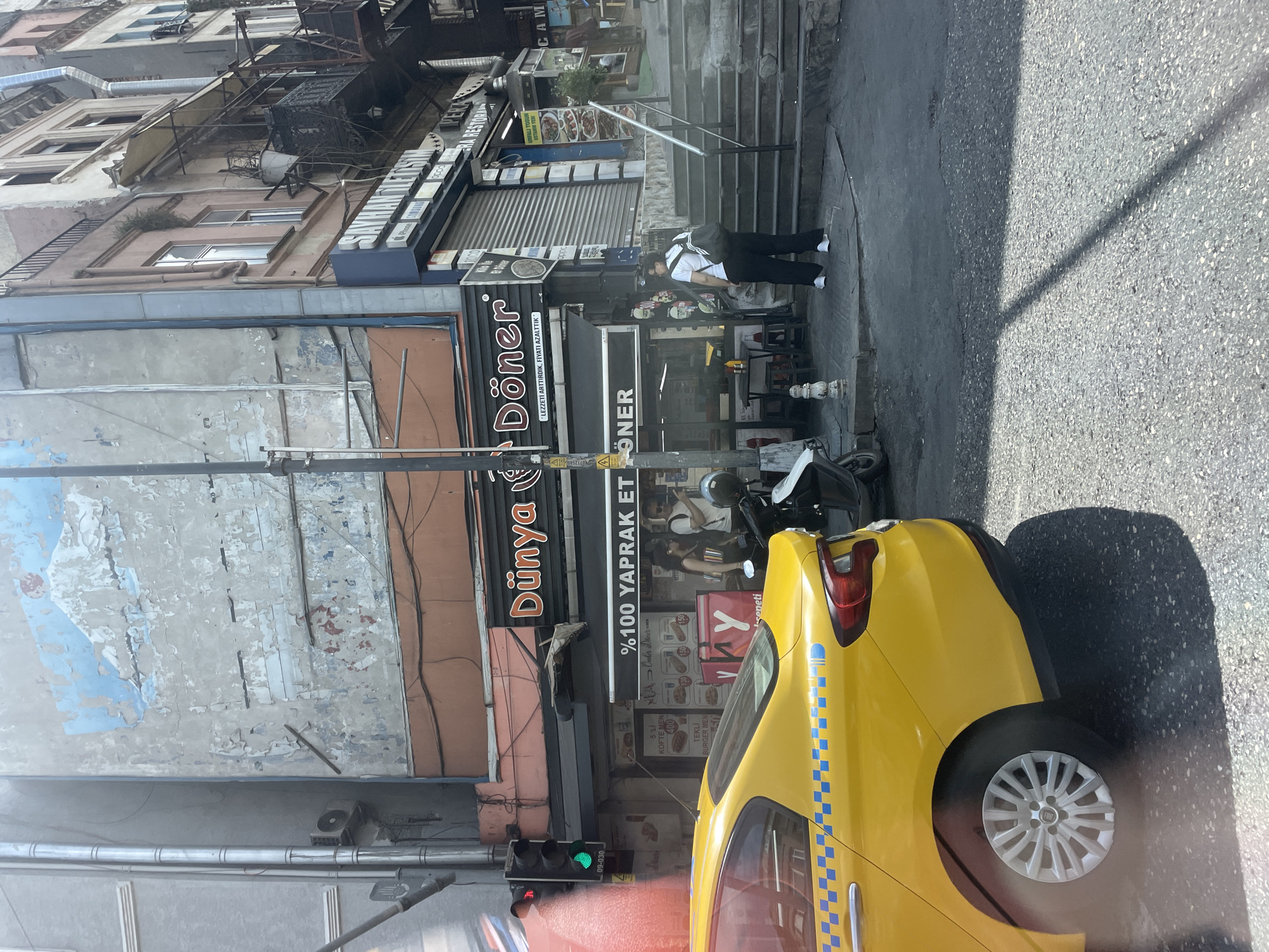This image shows a street scene in Beyoğlu, Turkey, likely in the Karaköy or Galata area, given the architecture and the presence of a döner shop. In the foreground, the rear quarter panel and wheel of a yellow taxi are visible, with a blue checkered stripe along its side. Behind the taxi, a street vendor establishment with the prominent signage "Dünya Döner" is displayed. The shop appears to be a typical döner kebab restaurant with outdoor seating. A traffic light is visible to the left of the döner shop, showing a green light. In the background, the buildings exhibit a mix of weathered facades, with peeling paint and visible pipes, characteristic of older urban districts. A set of outdoor stairs leads up to higher levels of the buildings on the right. A person wearing a white t-shirt and black pants, with a backpack, is walking up the stairs. Another individual is seen standing near the döner shop. The overall impression is of a busy, functional street in an established urban area.