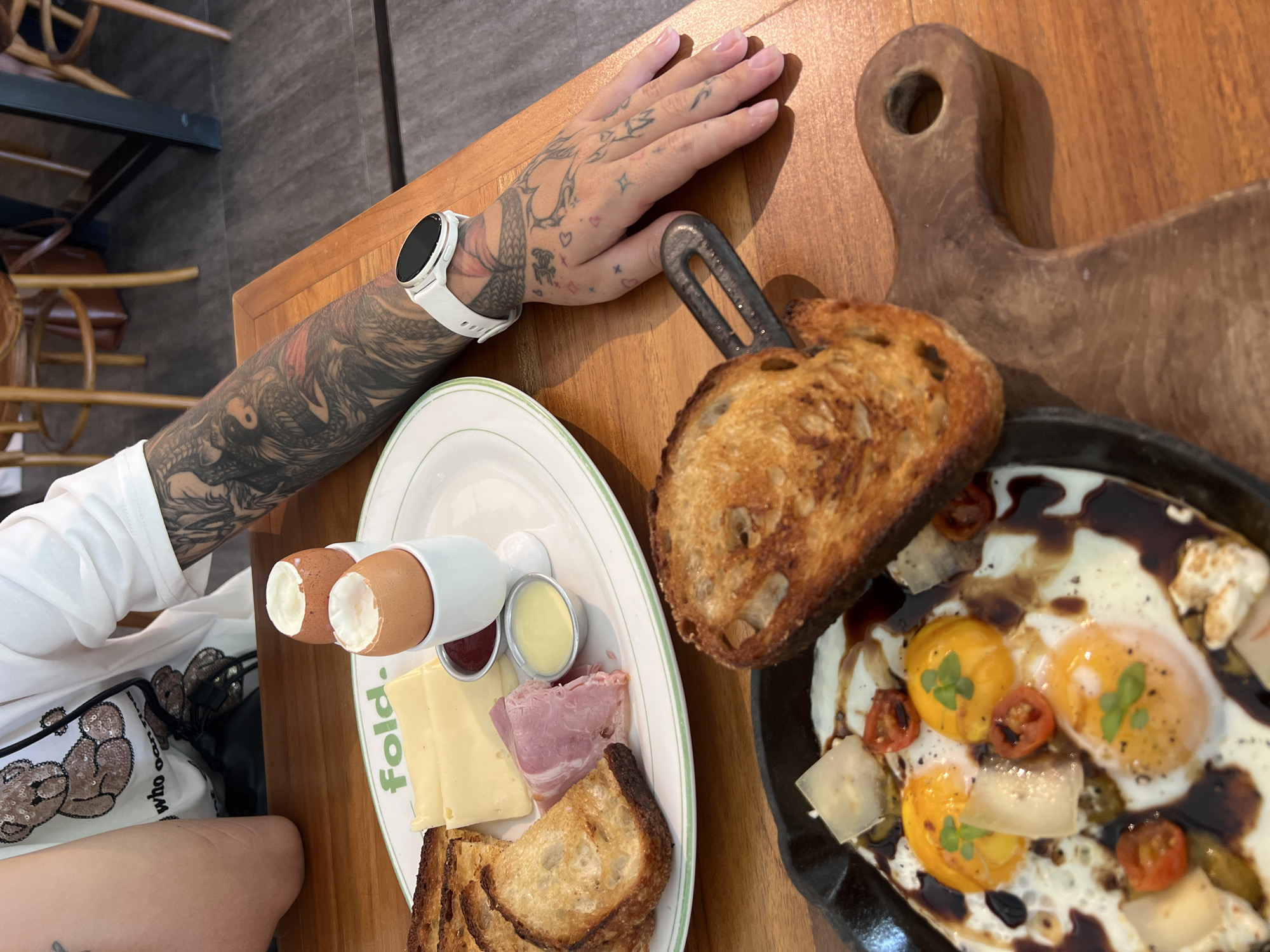 A top-down shot depicts a wooden table setting in what appears to be a restaurant in North Kuta, Indonesia, with a partially visible person with a heavily tattooed arm and hand positioned near the food. The individual wears a white wristwatch and a white shirt, and a shirt with a teddy bear design is visible. Two plates hold breakfast items. One is a white plate with a green rim displaying two soft-boiled eggs in egg cups, small containers of jam and what appears to be butter, slices of cheese and ham, and pieces of toast. Adjacent to this plate is a black skillet, on top of a wooden board, holding sunny-side-up eggs garnished with herbs and tomatoes and covered in a dark sauce. Slices of toasted bread lean against the skillet. The table is made of light-colored wood.