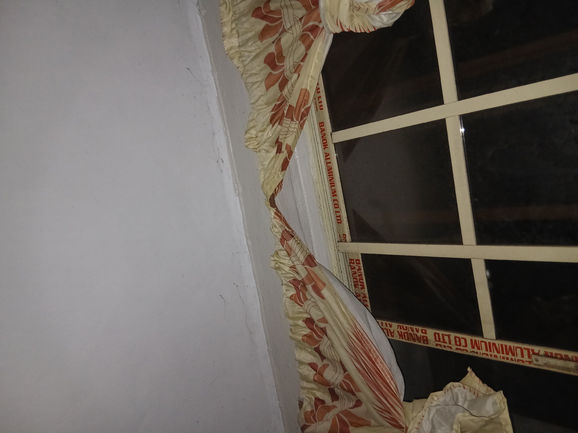 A close-up shot of a window with patterned curtains against a white wall. The window frame is made of aluminum. Text on the frame reads "VOR ALUMINIUM CO LTD BANDK AL," and "BANOK ALUMINIUM COLTO". The scene appears to be indoors.