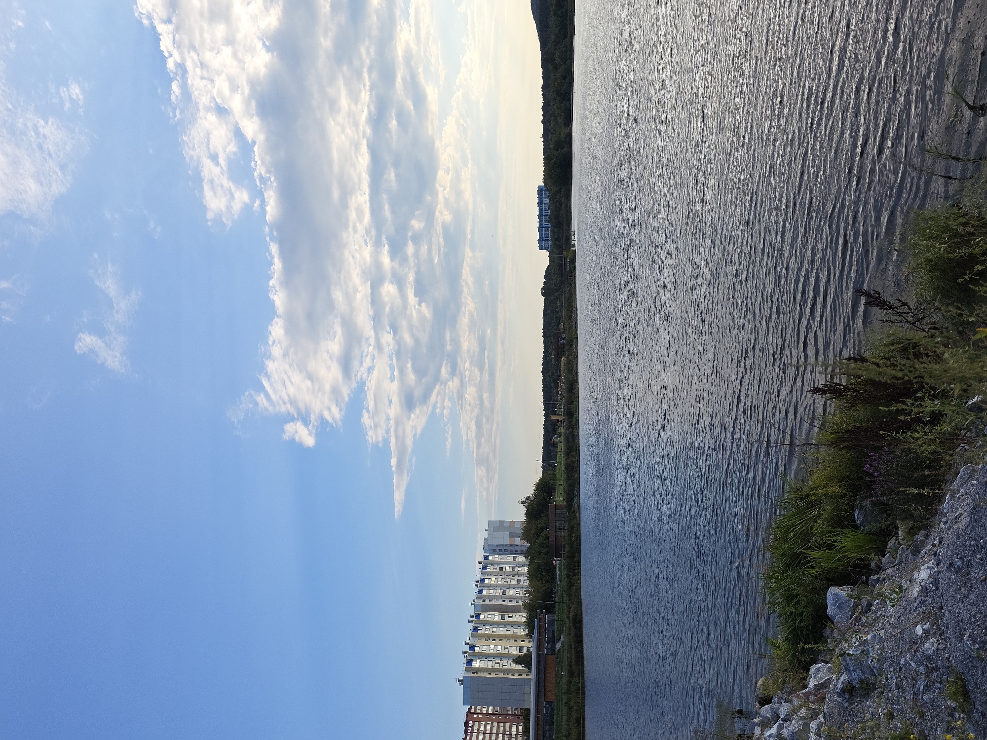 The image depicts a wide, calm body of water, likely a lake or reservoir, under a bright blue sky with scattered white clouds. In the background, a line of multi-story apartment buildings, characteristic of residential areas in Russia, stretches across the horizon on the left side. These buildings appear modern and are situated near the water's edge, separated by a strip of green trees and some smaller structures. On the right side of the image, the landscape transitions to a tree-covered hillside. The foreground shows a rocky, uneven shoreline with dense green vegetation growing along the water's edge. The water itself is rippled, suggesting a gentle breeze. The overall scene conveys a peaceful, urban-residential landscape by a natural water body, possibly located in or near the city of Miass, Russia, given the architectural style of the buildings.