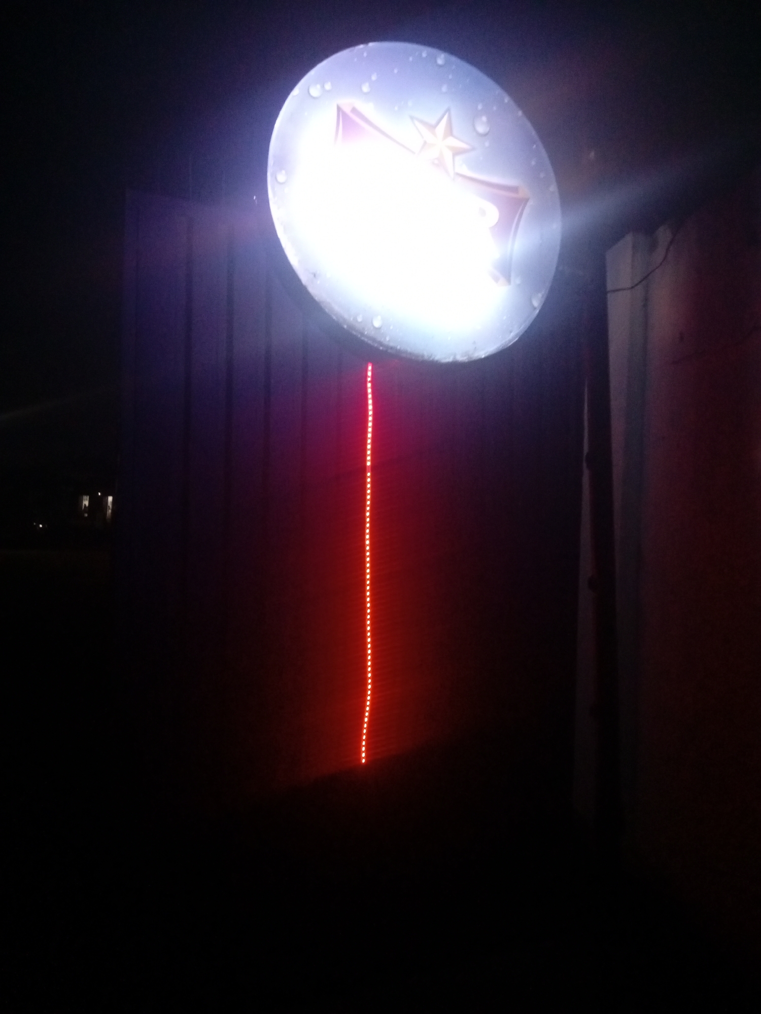 The image depicts a dimly lit scene, likely at night, in Sabon Gari, Nassarawa, Nigeria. A circular, illuminated sign is prominently featured, emitting a bright white light. The sign appears to have some texture or is possibly wet, with small circular droplets visible on its surface. A stylized star and a letter or symbol are faintly visible within the illuminated circle.
Extending downwards from the sign is a vertical line of small, bright red lights, resembling an LED strip. This red light casts a glow on the dark, corrugated metal surface behind it. The corrugated metal wall suggests a utilitarian structure, possibly a shop or a building facade.
To the left of the main illuminated sign, the background is dark, with faint lights visible in the distance, hinting at other structures or streetlights in the area. To the right of the sign, a vertical white element, possibly a pole or part of a building, is partially visible, and behind it, a darker, reddish surface can be observed. The overall impression is of a commercial or roadside establishment illuminated by its signage.