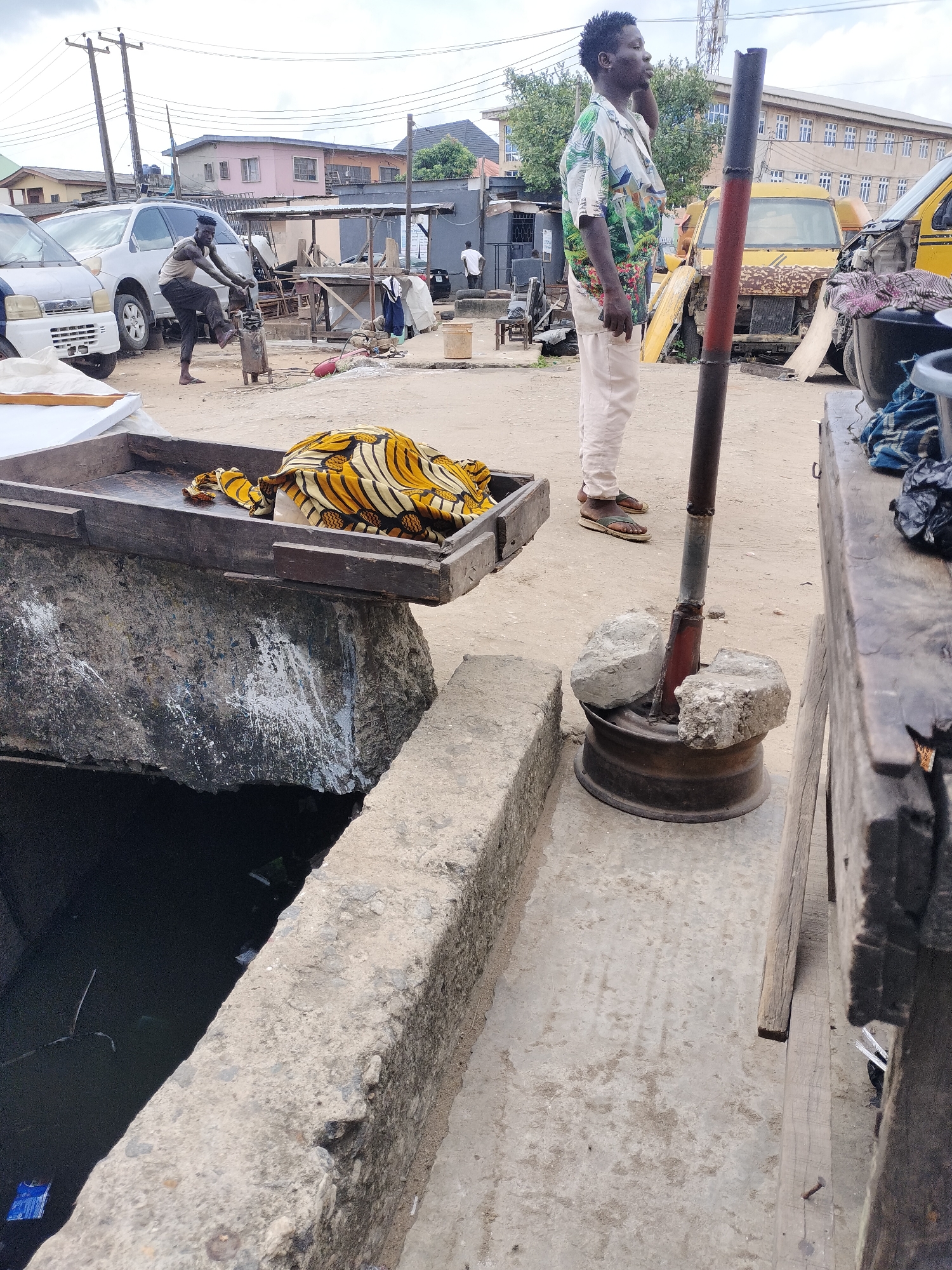 The image depicts a street scene in Lagos, Nigeria. A man in a floral shirt and light-colored pants stands near a makeshift structure of a metal pipe and wheel rim, stabilized by stones. To his left, a concrete structure borders a dark, water-filled channel. Atop the structure is a wooden tray containing folded yellow and white patterned fabric. In the background, another man is working a machine or tool near a white van. In the distance, buildings can be seen under a cloudy sky. A yellow van is also visible to the right of the man in the floral shirt. Overall, the scene has a workaday, somewhat disorganized atmosphere.