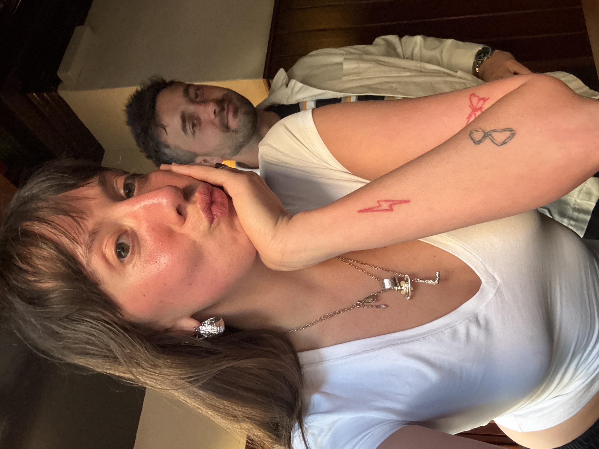 A medium-shot, rotated slightly counter-clockwise, shows a selfie-style image of a woman and a man indoors.
The woman is in the foreground. She has medium-length brown hair with bangs, light skin, and is making a kissing face towards the camera. She wears a white t-shirt and a silver necklace with a pendant. On her right arm, visible due to the short sleeves, there are three small tattoos: a lightning bolt, a pair of glasses, and a bow. An earring is visible in her left ear.
The man is in the background, partially obscured. He has short dark hair with some graying, a beard, and light skin. He is wearing a white shirt or jacket with a dark-colored stripe. A watch is visible on his left wrist.
They are both lying down, possibly in bed. The background is dark, with wooden paneling. The setting seems to be a bedroom, possibly in a hotel or home.
No location in the city of London, United Kingdom is possible to derive.