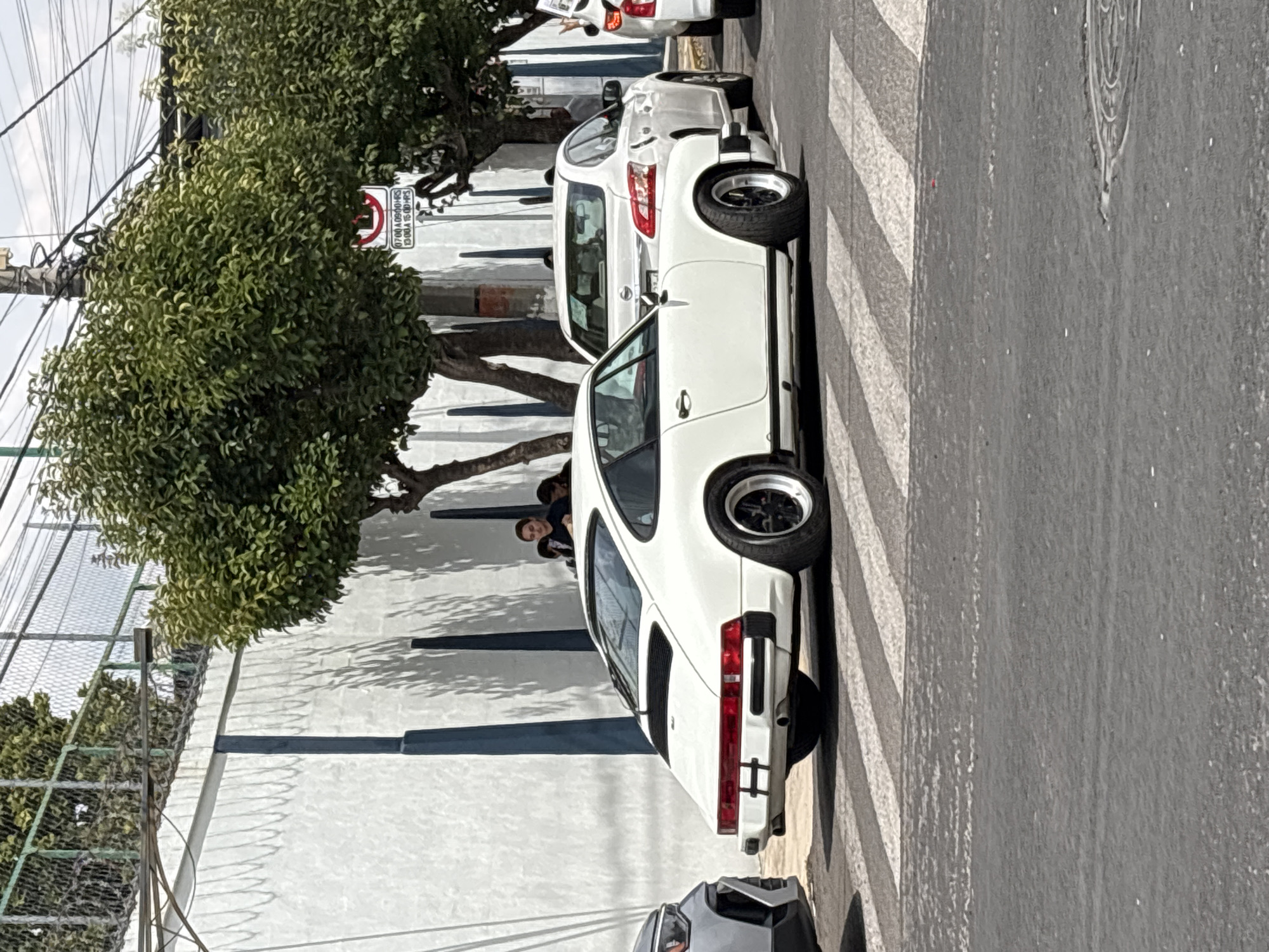 The image shows a white Porsche parked on a street, likely in the city of Puebla, Mexico, based on the style of the architecture and signage visible. The car is parked next to a pedestrian crosswalk. Behind the car, another white vehicle is parked. A white wall runs parallel to the street, and a tree with dense foliage partially obscures a 'no parking' sign, which is displayed from 07:00 to 09:30 and from 13:00 to 15:00. Two people are standing near the base of the tree, possibly interacting with each other. Overhead, there's a web of electrical cables and a wire fence. Other vehicles appear to be parked at either end of the frame.