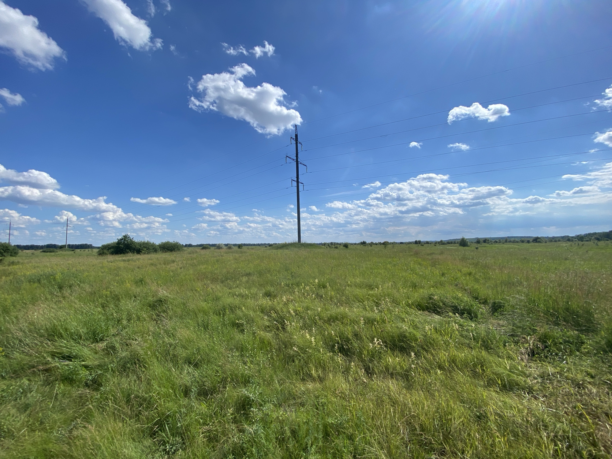 A wide-angle, eye-level, full shot captures a bright, sunny day over a grassy field in Matiashivka, Ukraine. The sky is a vibrant blue, dotted with fluffy white clouds. A solitary, dark electrical tower stands tall in the middle ground, supporting multiple parallel wires that stretch across the frame. The field is covered in tall, green grass, with some yellowing stalks and scattered wildflowers visible. In the distance, the horizon is lined with a few trees and bushes, with more of the electrical transmission line visible. The sun glints brightly in the upper right corner of the image, creating lens flare. There are no people or animals visible in the scene.
