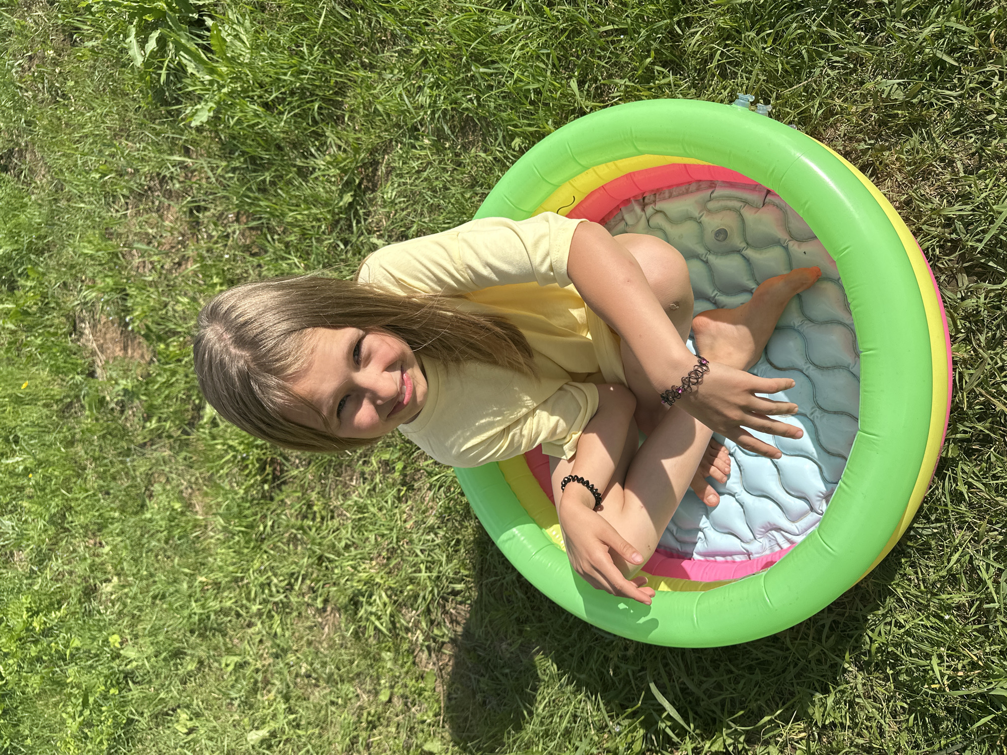 A young girl with blonde hair is sitting in a small, colorful inflatable pool in a grassy area. She is wearing a yellow t-shirt. The pool has green, yellow, and pink sections around its rim, and the interior is light blue with a quilted pattern. Her legs are bent, and her feet are visible inside the pool. She is looking towards the camera with a smile. Two dark bracelets are visible on her wrists. The scene appears to be outdoors on a sunny day, with green grass surrounding the pool. The sunlight casts shadows on the grass, suggesting it is midday. The presence of the inflatable pool and the sunny weather indicates a recreational activity, likely during warmer months. The location is identified as Mokino, Russia. There is no visible text in the image.