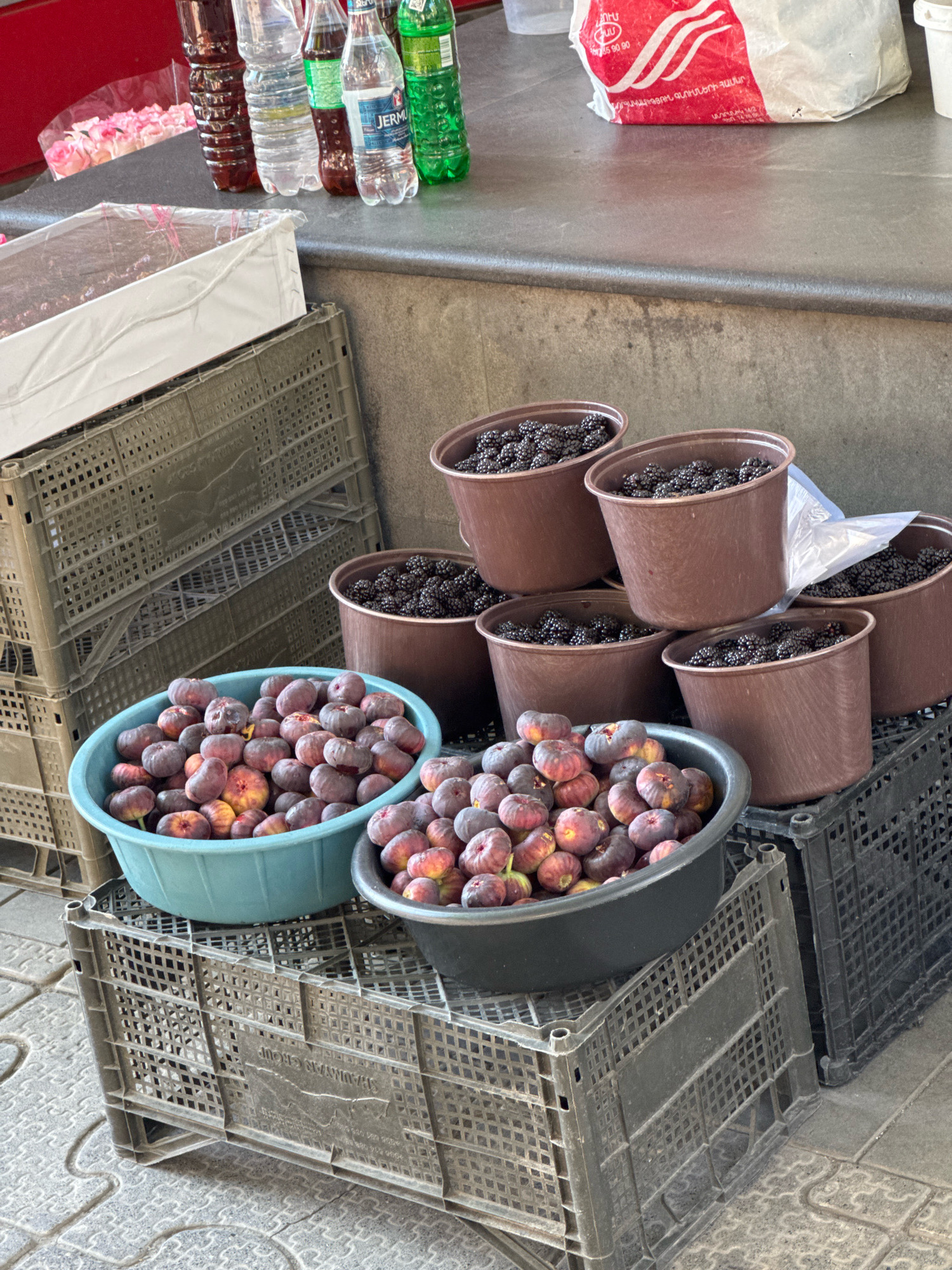 The image depicts a street market scene in Yerevan, Armenia. In the foreground, a light blue bowl is filled with ripe figs. To the right of the figs, a dark gray bowl also contains figs. Behind these bowls are several brown plastic buckets brimming with blackberries. These containers are stacked on and around various plastic crates, some dark gray and others black. In the background, to the left, a white box is covered with clear plastic. Further back and to the left, several plastic bottles are visible, including a dark reddish-brown liquid, clear water, and green beverages. Behind the bottles, a partially visible bag with red and white branding sits on a grey countertop. The ground appears to be paved with patterned tiles. The overall setting suggests an outdoor market stall, likely a fruit vendor.
