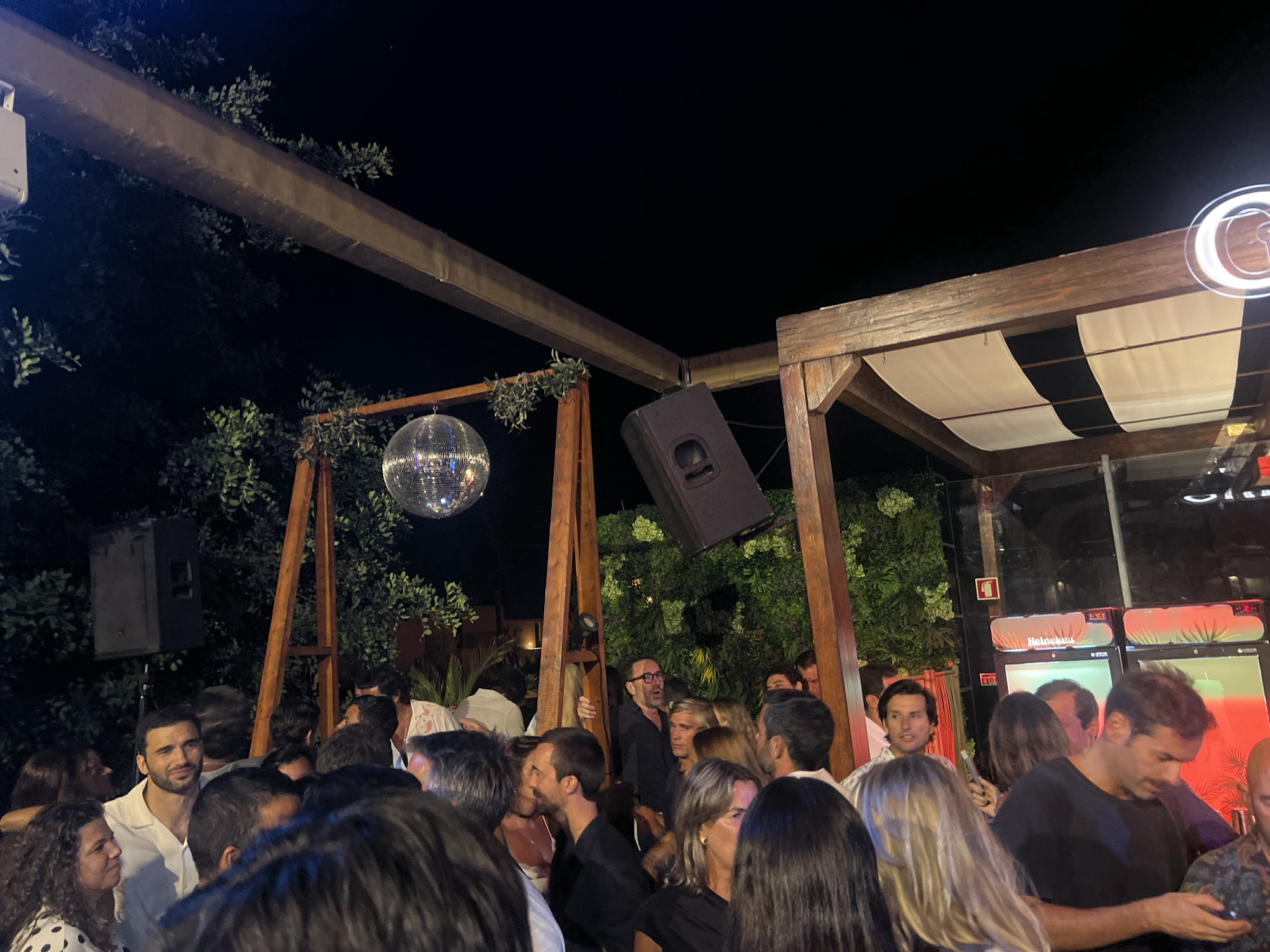 The image depicts a nighttime gathering of people at an outdoor venue in Quarteira, Portugal. A disco ball hangs overhead, reflecting light. A large speaker is mounted on a wooden structure. The crowd is densely packed, with individuals engaged in conversation and socializing. The background features foliage and wooden architectural elements, suggesting an open-air bar or club. Several people are visible in the foreground and midground, some looking towards the camera. The scene is illuminated by artificial lighting, contributing to a festive atmosphere.