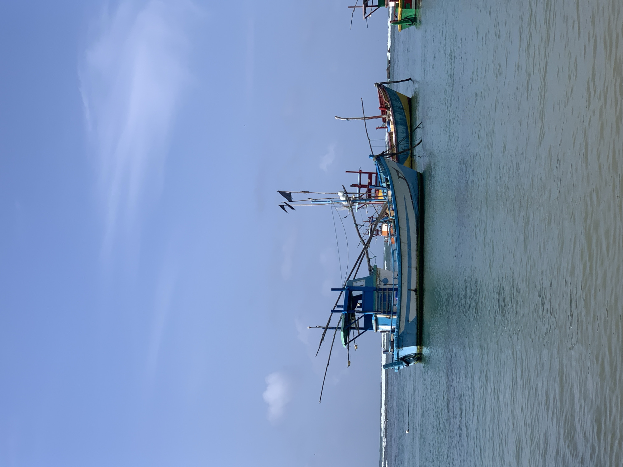 The image captures a serene coastal scene in Mirissa, Sri Lanka. In the foreground, calm turquoise waters gently lap against the shore. Several fishing boats are anchored offshore, their colorful hulls a vibrant contrast against the blue of the sea and sky.
The most prominent boat is a blue and white vessel with a sturdy structure, equipped with fishing rods extending outwards. Two black flags fly from its mast, adding a dramatic touch to the scene. Behind it, a yellow boat with a red trim is also visible, its lines simpler, suggesting a different purpose or design. Further to the right, a glimpse of a green and yellow boat hints at a fleet of vessels.
The background reveals a line of breaking waves, indicating the presence of a reef or shallow waters further out. Beyond the water, a clear blue sky stretches across the horizon, with a few wispy clouds adding texture to the expanse. The overall atmosphere is peaceful and tranquil, showcasing a typical day for the fishing community in Mirissa. There are no people visible in the frame, giving the scene a sense of quiet solitude. The overall impression is one of a picturesque fishing harbor, bathed in bright daylight.