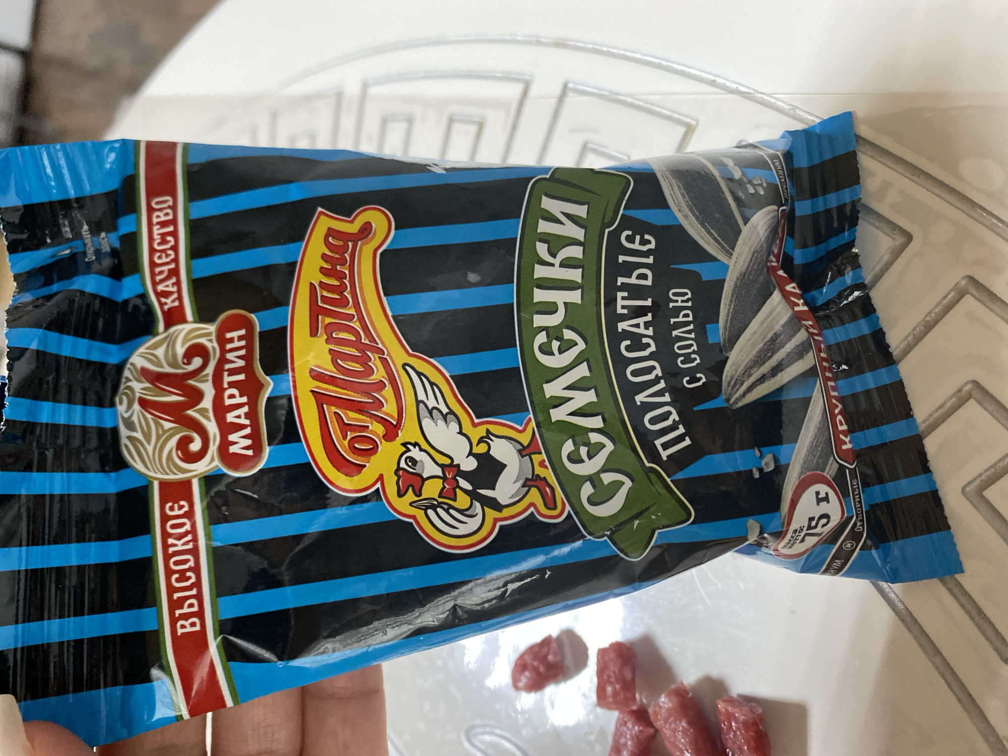 The image shows a close-up of a packaged product, likely sunflower seeds, with Russian text. The packaging is predominantly blue and black striped with red and yellow accents. Prominently displayed are the brand name "МАРТИН" (MARTIN) and the product name "СЕМЕЧКИ ПОЛОСАТЫЕ С СОЛЬЮ" (Striped Sunflower Seeds with Salt). There is also a graphic of a chicken wearing a bowtie and holding a utensil. A small red circular label indicates "75 г" (75g). The product is partially obscuring a white surface, possibly a table, and a metal rack. Several small pieces of red food, resembling cured meat, are scattered on the white surface to the left of the packaging. The image appears to be taken indoors. No specific location in the city of Chirsha-Tartysh, Russia, can be identified from the visual information provided.