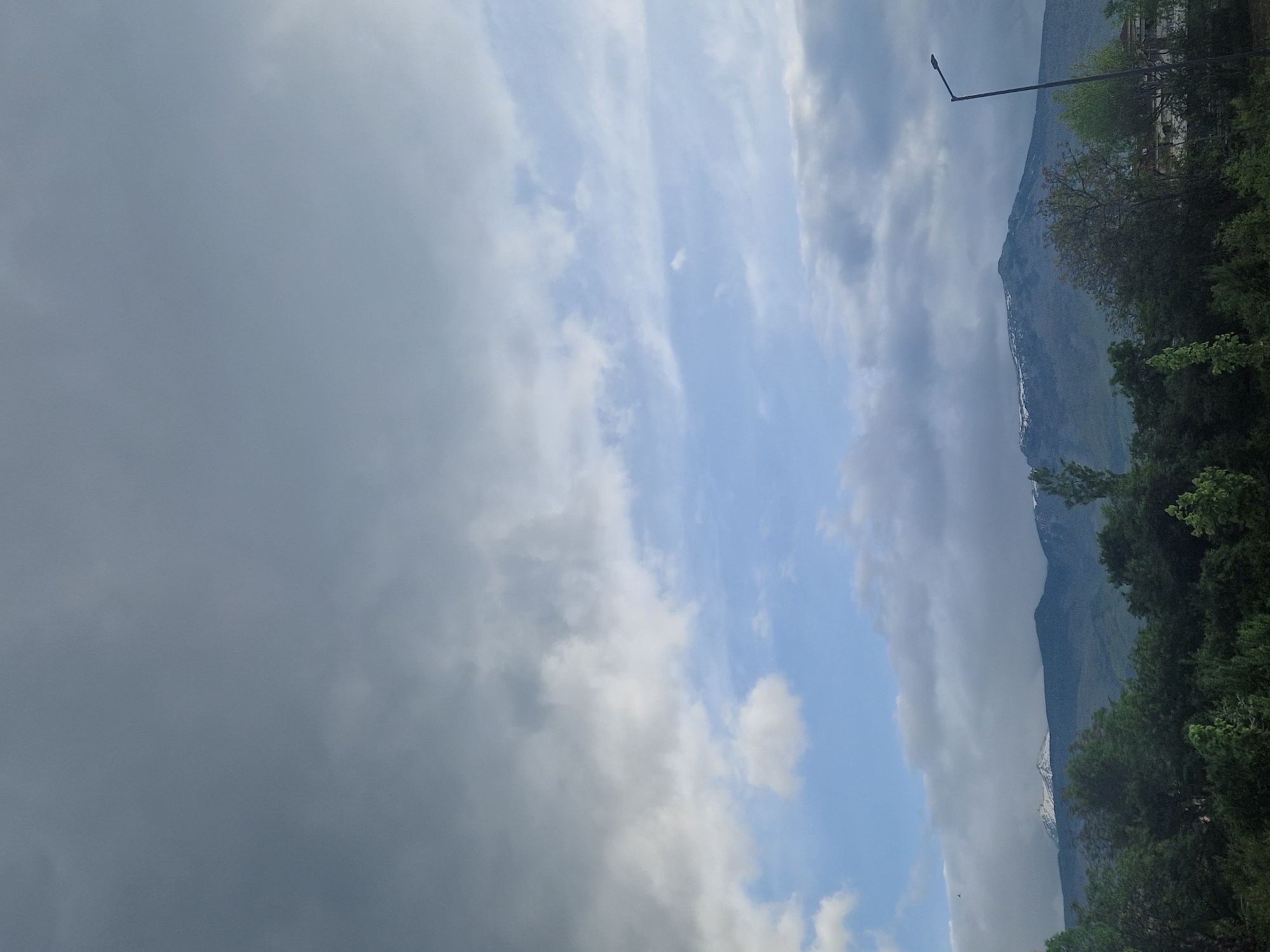 The image depicts a view of a mountain range partially obscured by heavy, dark clouds on the left side. There are also patches of lighter, white clouds and some blue sky visible. Trees are prominent along the base of the mountain, with a street light seen amongst them. The location is in the city of Unknown, Unknown.