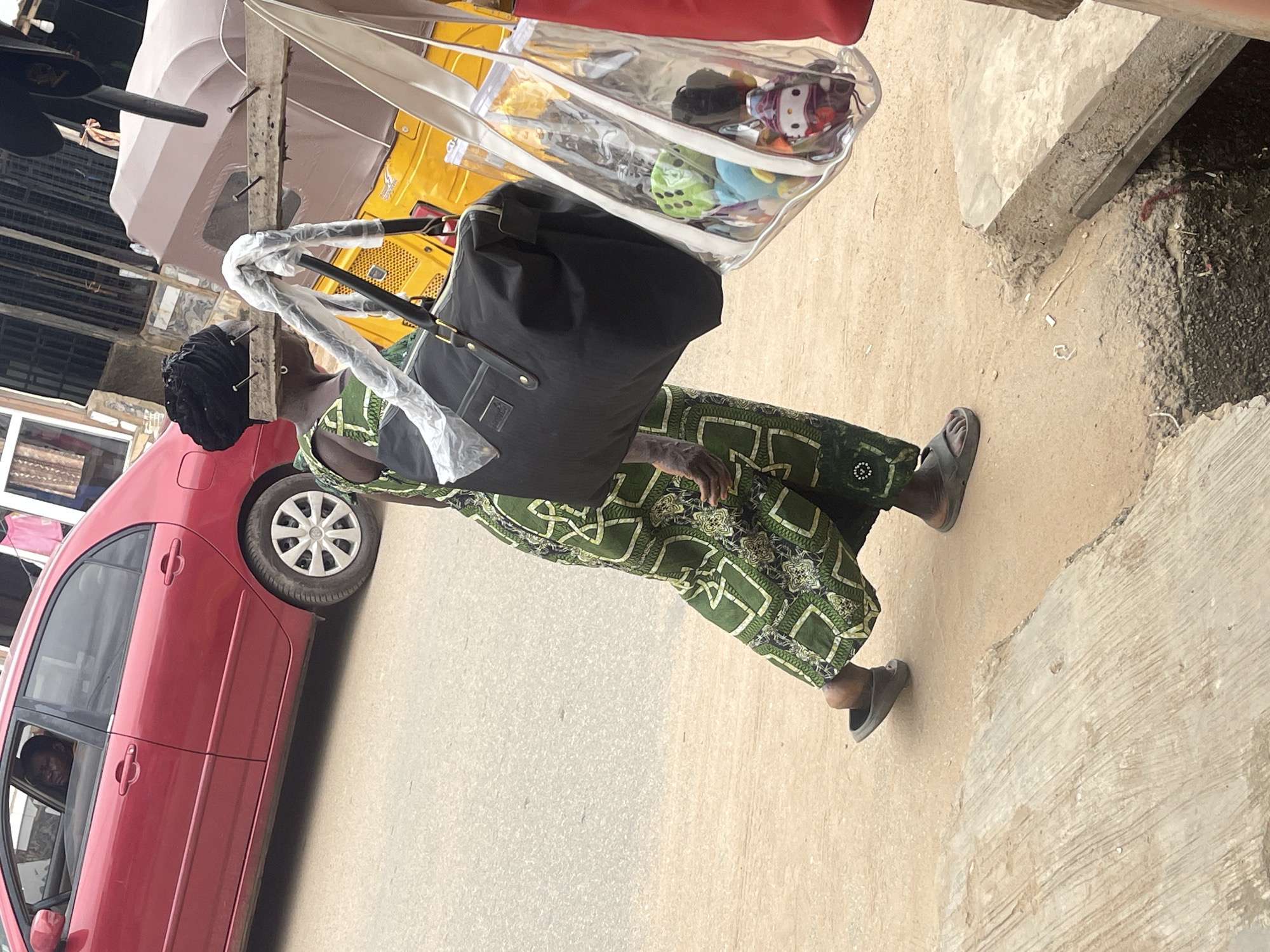 A woman is walking on a dirt and paved surface in front of a red sedan. She is wearing a green patterned dress and dark sandals. She is carrying a large black duffel bag over her shoulder and a clear plastic tote bag filled with stuffed toys. In the background, a yellow trishaw is parked, and a red sedan is visible with a person inside. The environment appears to be an outdoor market or roadside stall, likely in Abuja, Nigeria, given the presence of the trishaw, common mode of transport in West Africa.