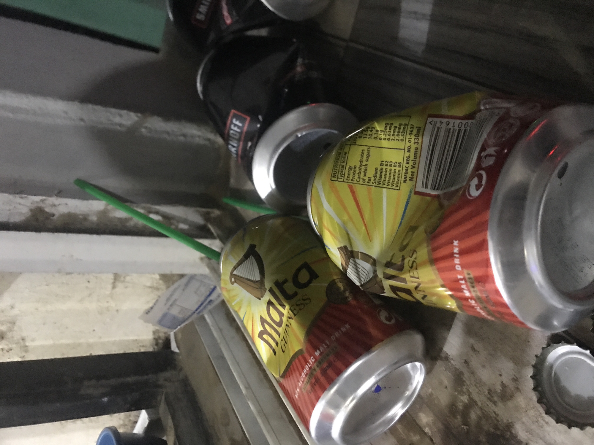 In New Stadium, Nigeria, on a worn, wooden surface, lie several empty beverage cans. The cans are a mix of brands and colors, with two prominent Malta Guinness cans in the foreground. These cans are adorned with a distinctive gold and red design featuring the Malta Guinness logo. One of the Malta Guinness cans has a straw inserted, a bright green one. In the background, a dark can, possibly a Smirnoff, is partially visible. The scene appears to be outdoors, with a concrete wall in the background and hints of natural light. The overall impression is one of casual disposal, with the cans scattered as if recently consumed and left behind.