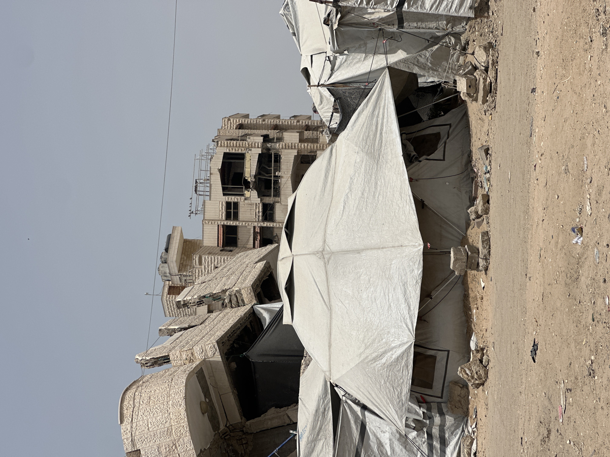 The image shows a row of white tents erected next to a damaged building. The building is multiple stories tall with significant structural damage, including missing walls and scaffolding on one side. The tents appear to be makeshift shelters, possibly for displaced people. The ground is dirt and debris. The location is in a city where a building has been damaged.