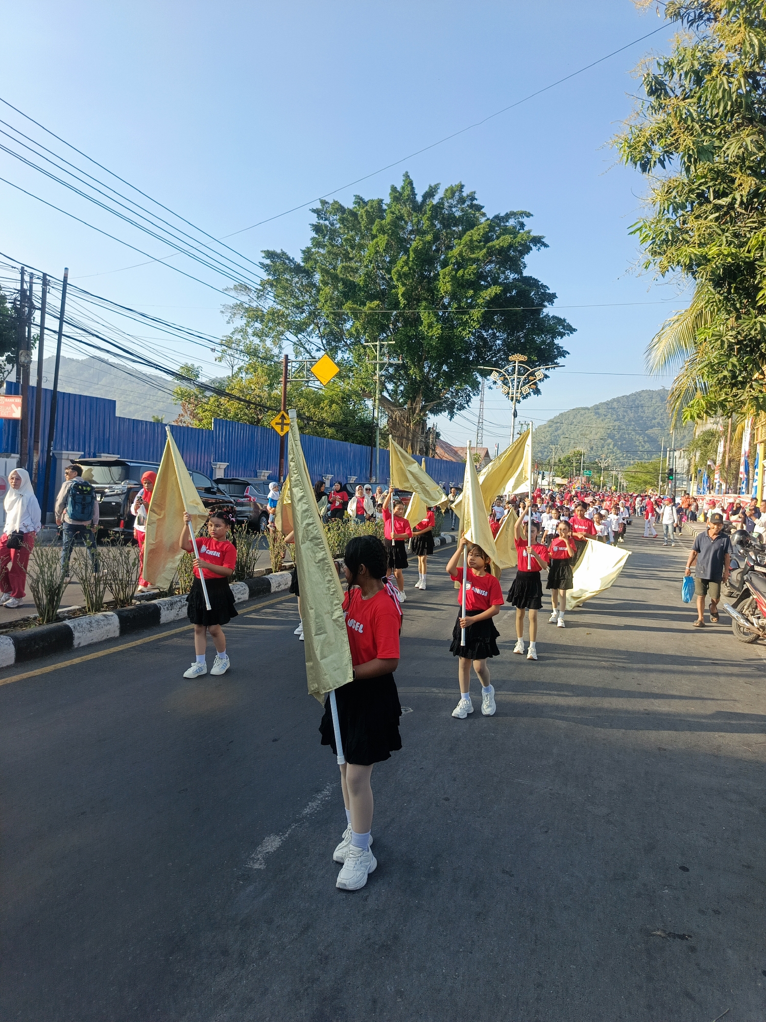 A group of young girls, dressed in red shirts and black skirts, are participating in a parade on a street in Gorontalo, Indonesia. They are holding large, golden flags. The parade appears to be taking place on a sunny day, with clear blue skies. The street is lined with trees and buildings, and there are spectators on the sidewalks. In the background, mountains can be seen. The overall scene suggests a festive and lively atmosphere.